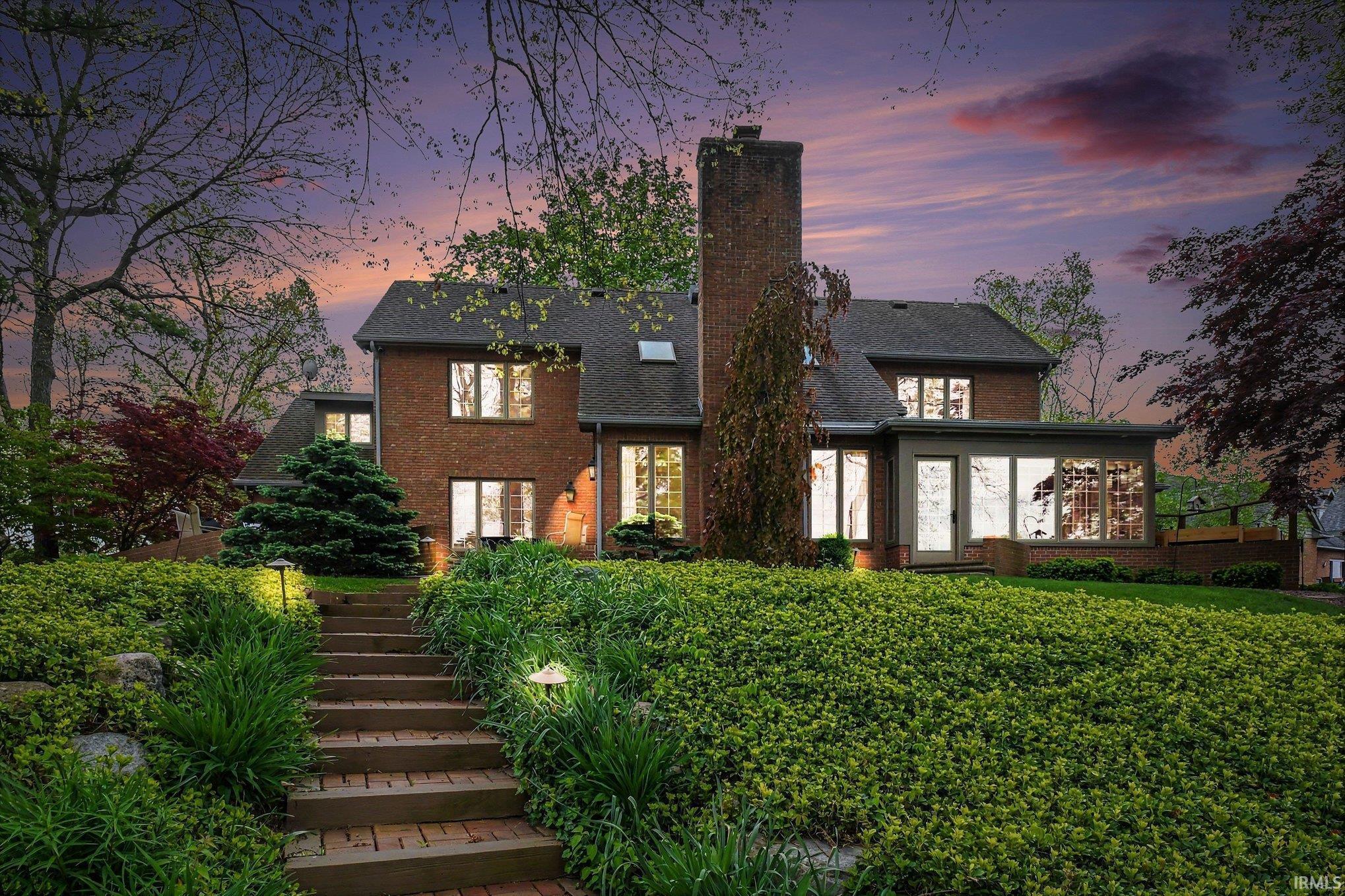 Brick exterior featuring a multi-level roofline, extensive landscaping, a sunroom with large windows, and an illuminated brick pathway