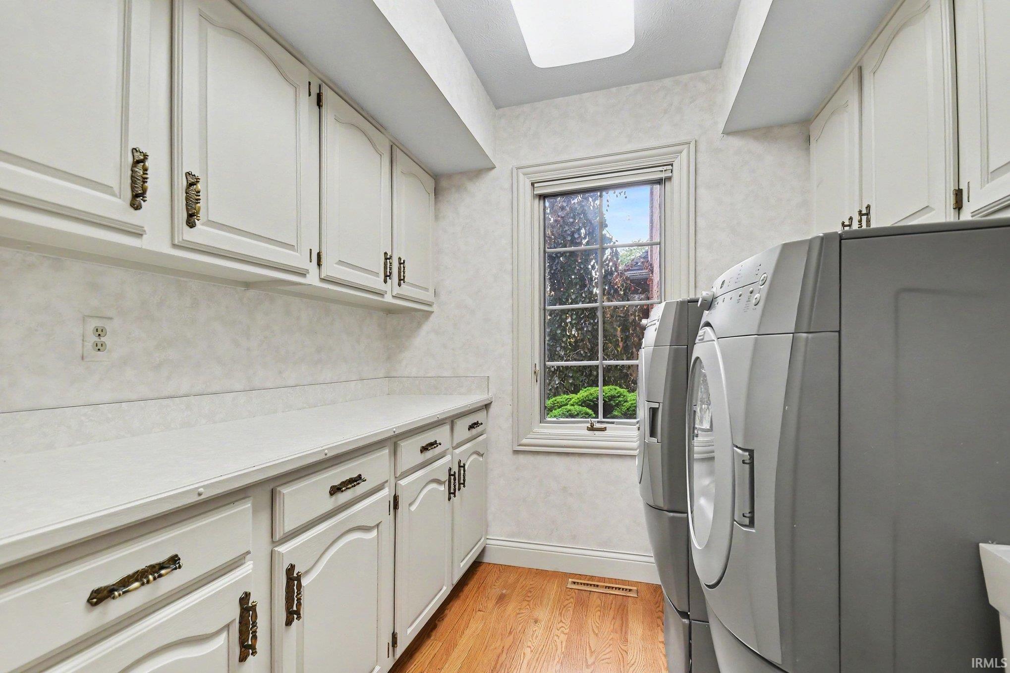 Laundry area featuring light wood-finish flooring, white cabinetry with decorative hardware, extensive countertop space, and a window providing natural light