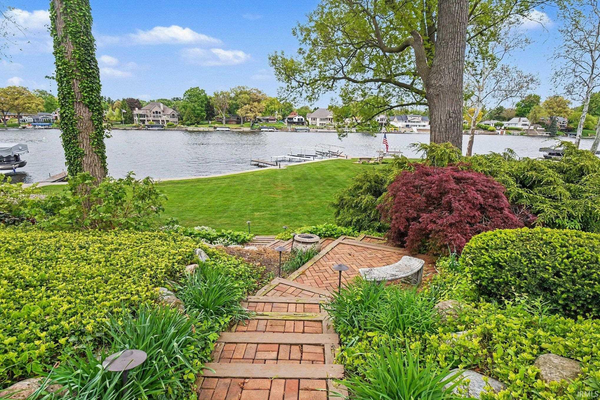 Landscaped backyard featuring brick paver steps, mature trees, diverse plantings, a stone bench, and expansive lawn leading to the waterfront