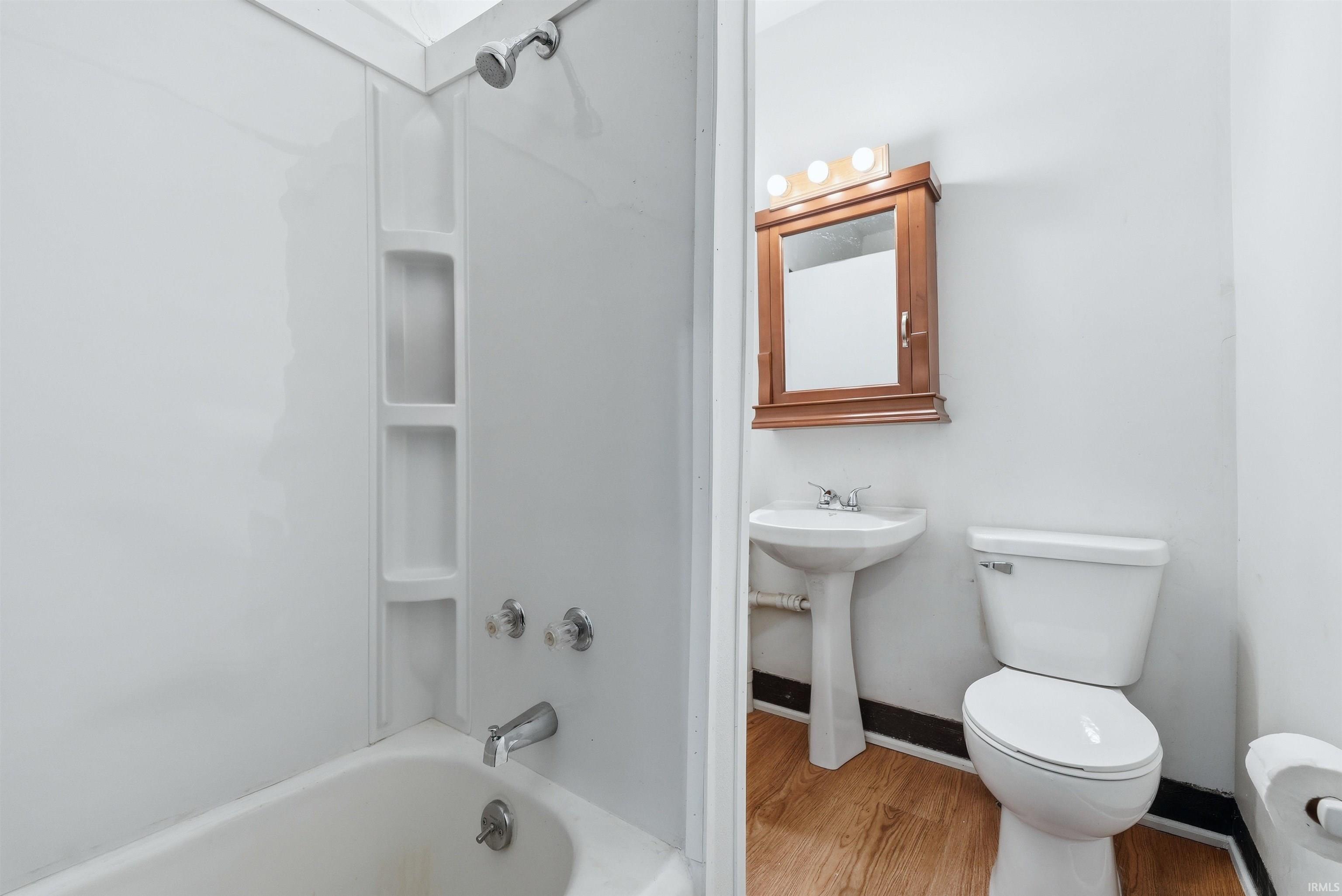 Bathroom featuring a white tub with integrated shelving, a pedestal sink, a toilet, a wood-finish medicine cabinet, and wood-finish flooring