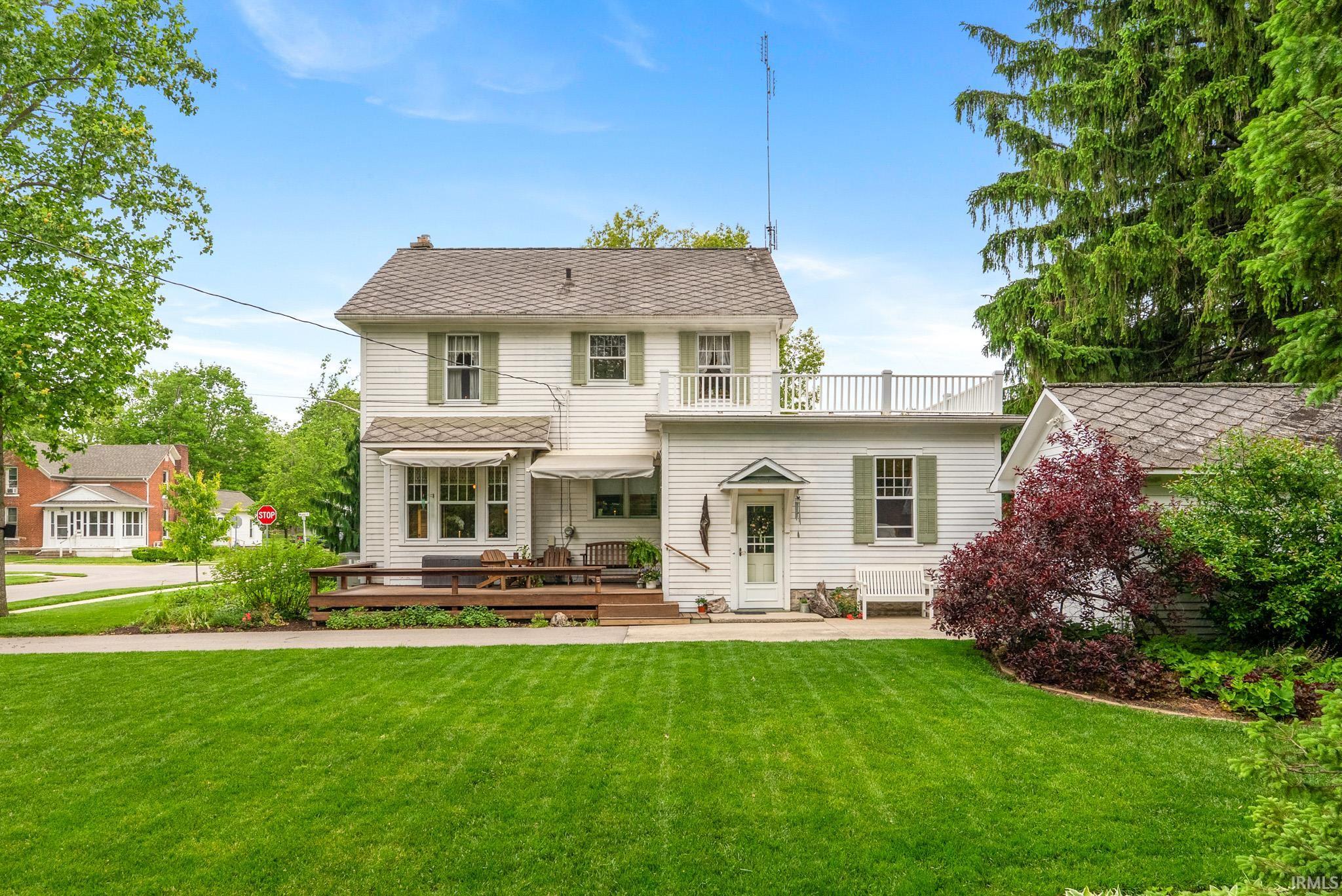 White siding residence featuring a spacious lawn, wood deck, second-story balcony, and mature landscaping