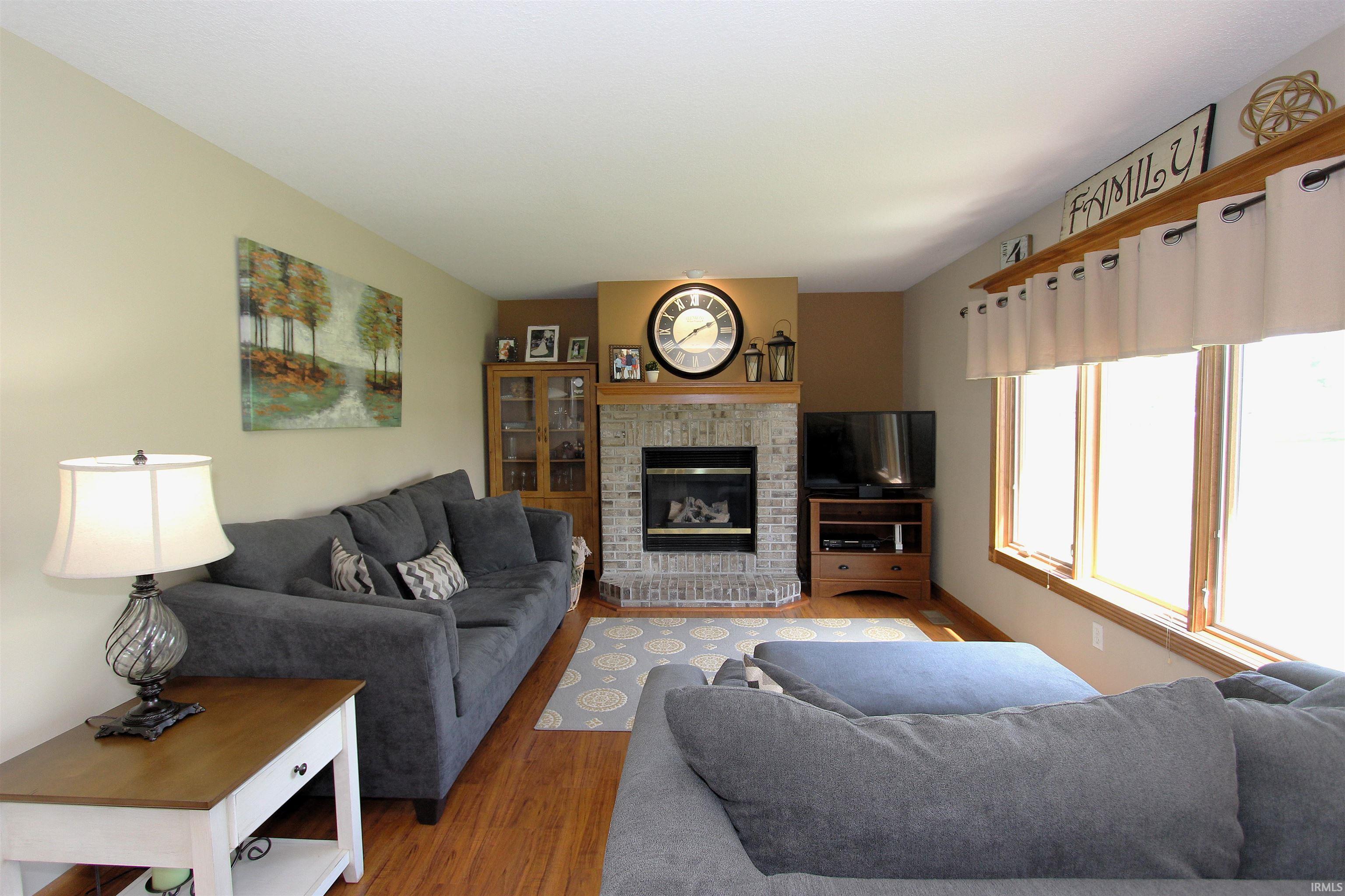 Spacious living area featuring warm wood-finish flooring, a prominent brick fireplace with a mantel, and multiple large windows with wood trim