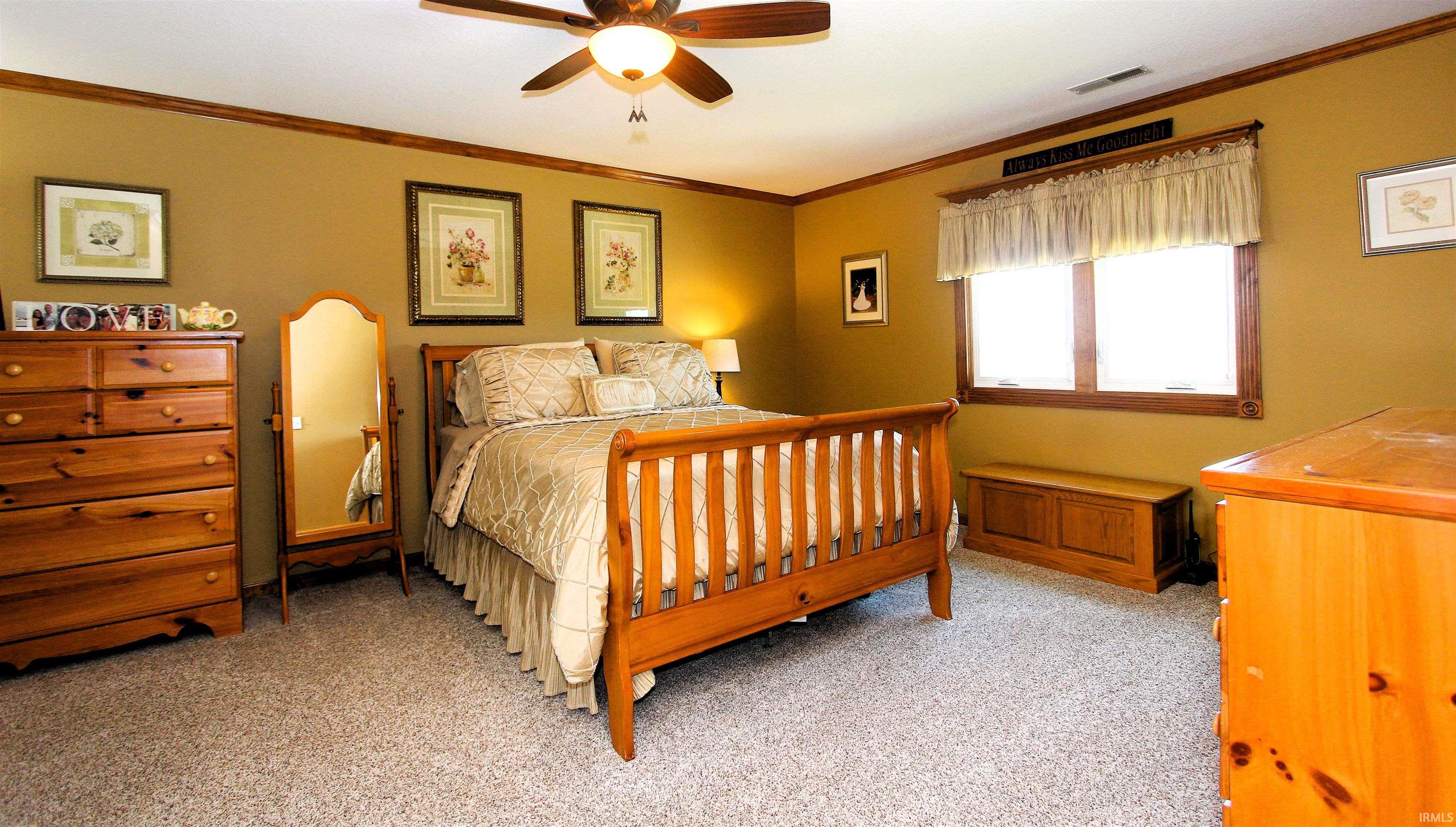 Carpeted bedroom featuring a ceiling fan with integrated lighting, a broad window with wood trim, and crown molding