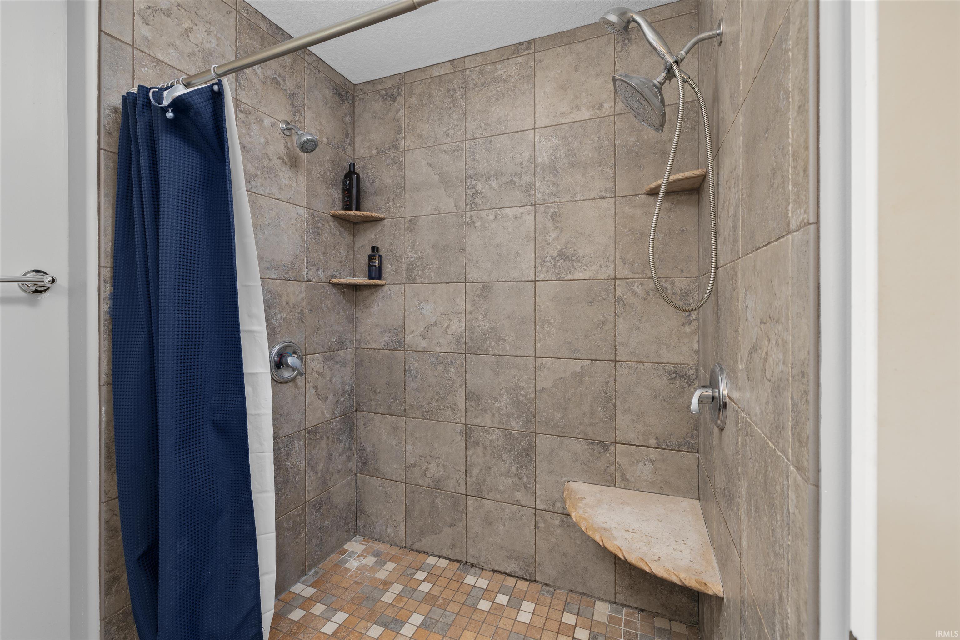 Shower enclosure featuring large format stone-look wall tiles, mosaic tile flooring, a built-in corner bench, and a dual-head shower system