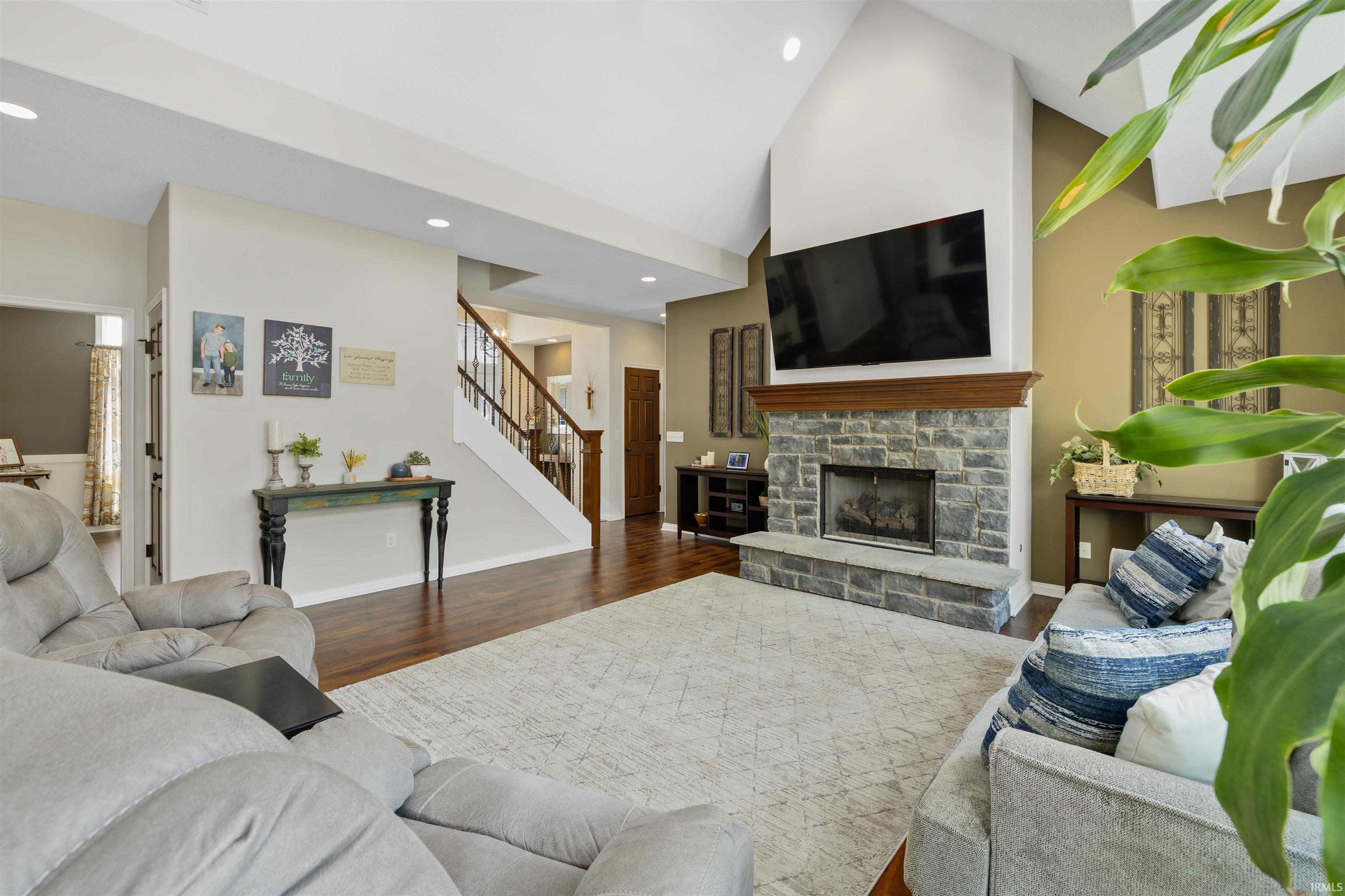 Living area featuring a stone fireplace with a wood mantle, vaulted ceilings, recessed lighting, wood-finish flooring, and a visible staircase with wood railing