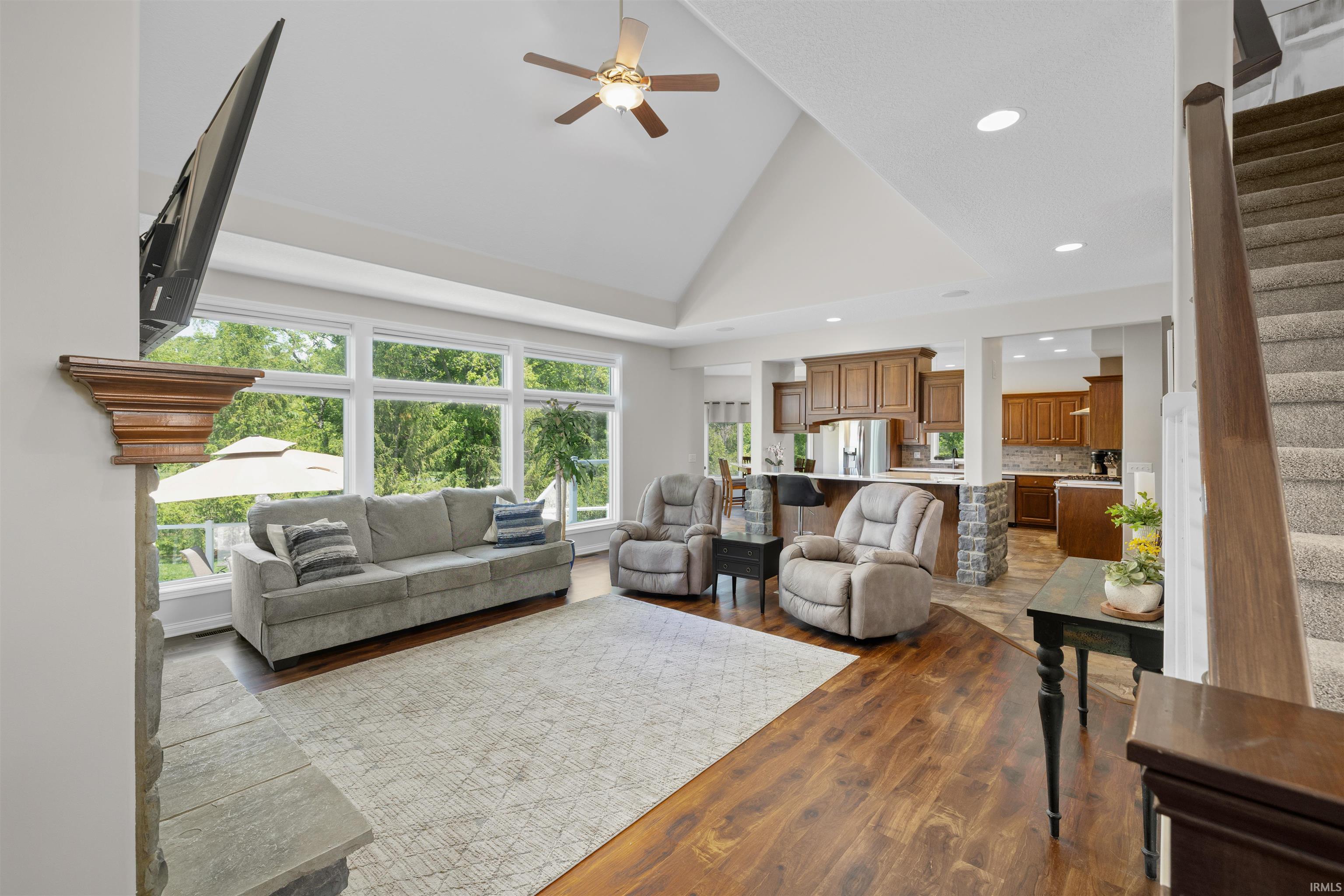 Spacious living area featuring wood-finish flooring, a vaulted ceiling with a fan, and large windows overlooking a wooded setting