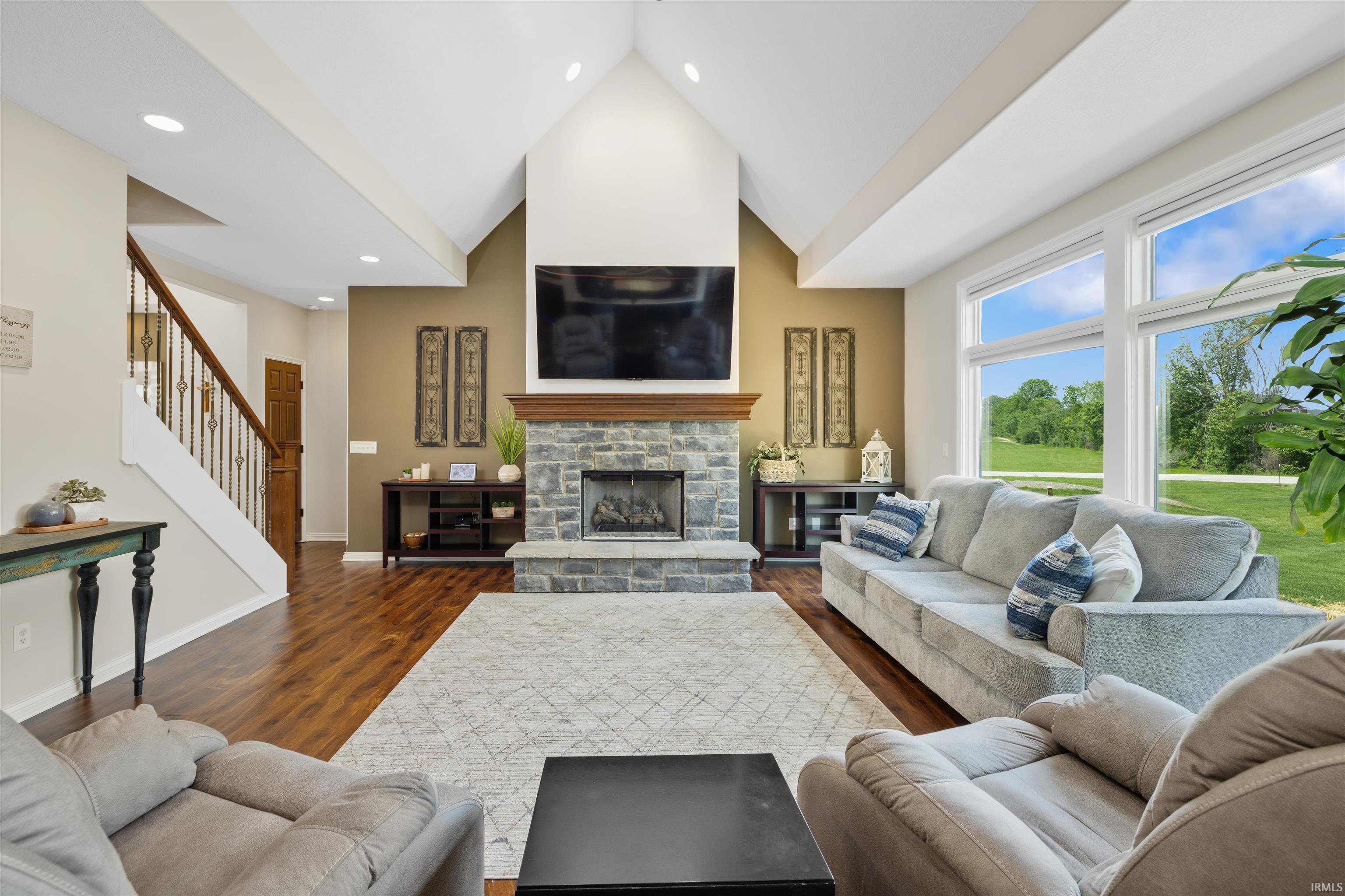 Vaulted ceiling living area featuring a stone-clad fireplace with wood mantel, large picture windows, wood-finish flooring, and a staircase with an iron balustrade