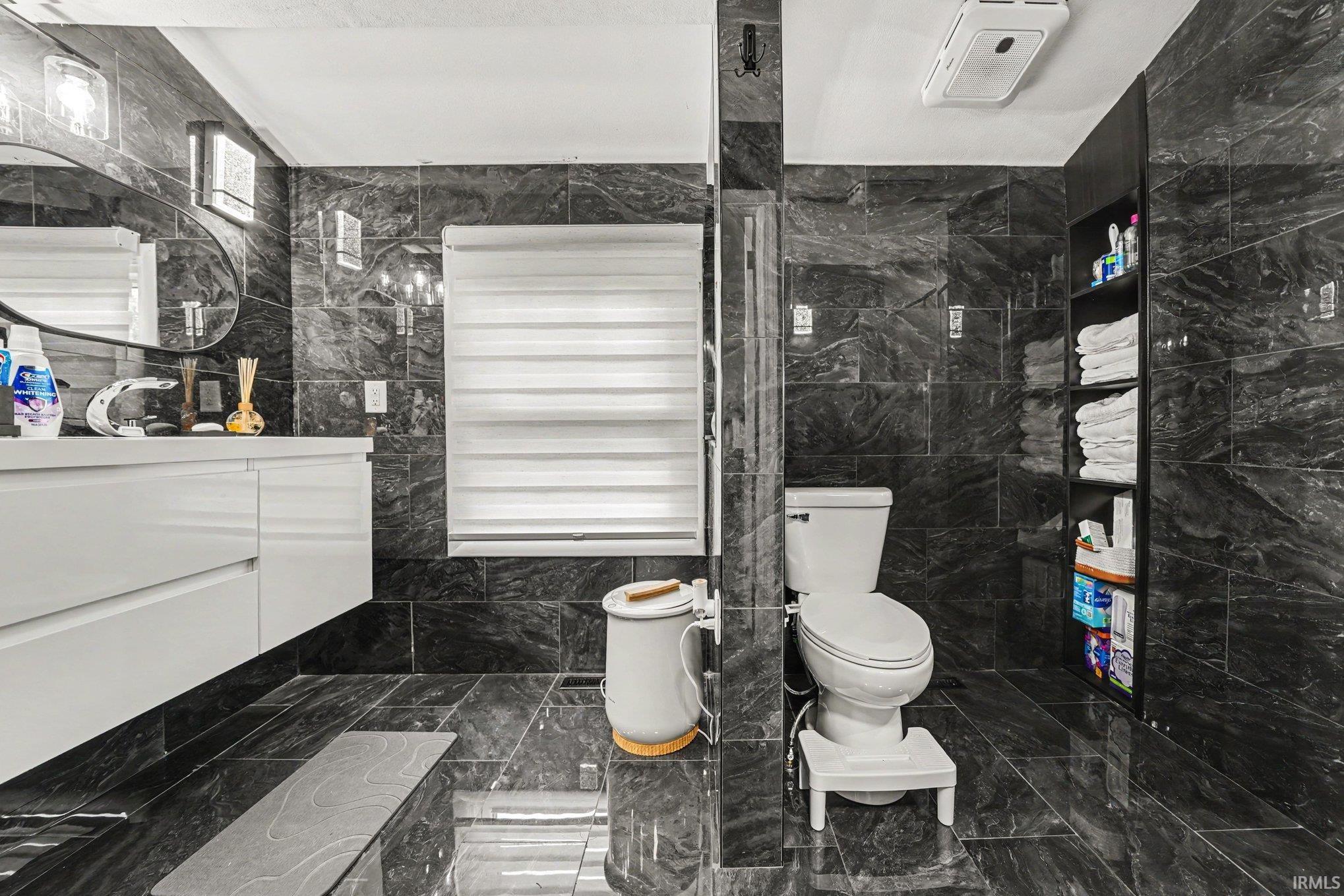Bathroom featuring dark stone-finish wall and floor tile, a floating white vanity with a chrome faucet, and a toilet with an adjacent built-in shelving unit