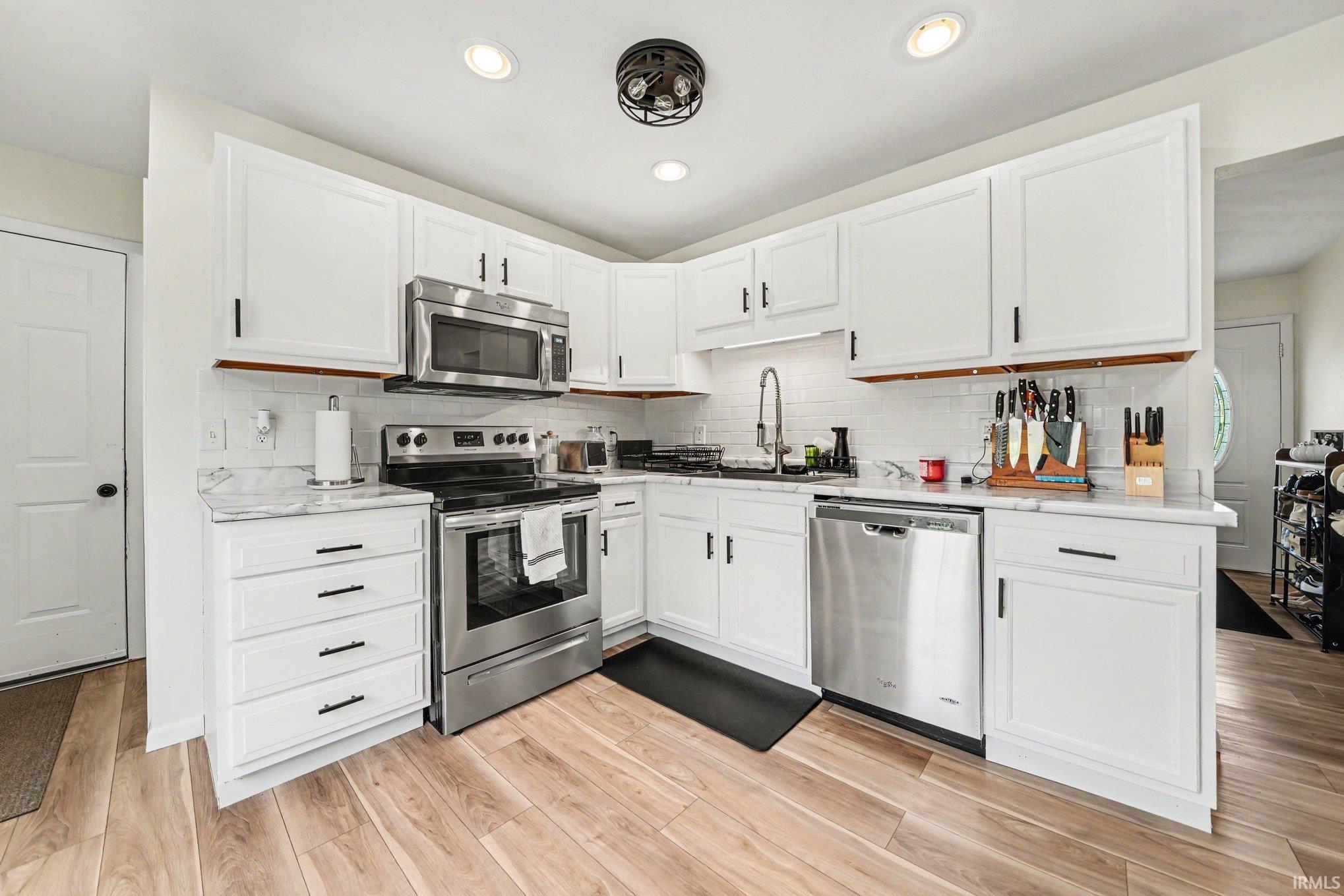 Updated kitchen featuring white shaker-style cabinetry, stainless steel appliances, marble-look countertops, subway tile backsplash, and light wood-finish flooring