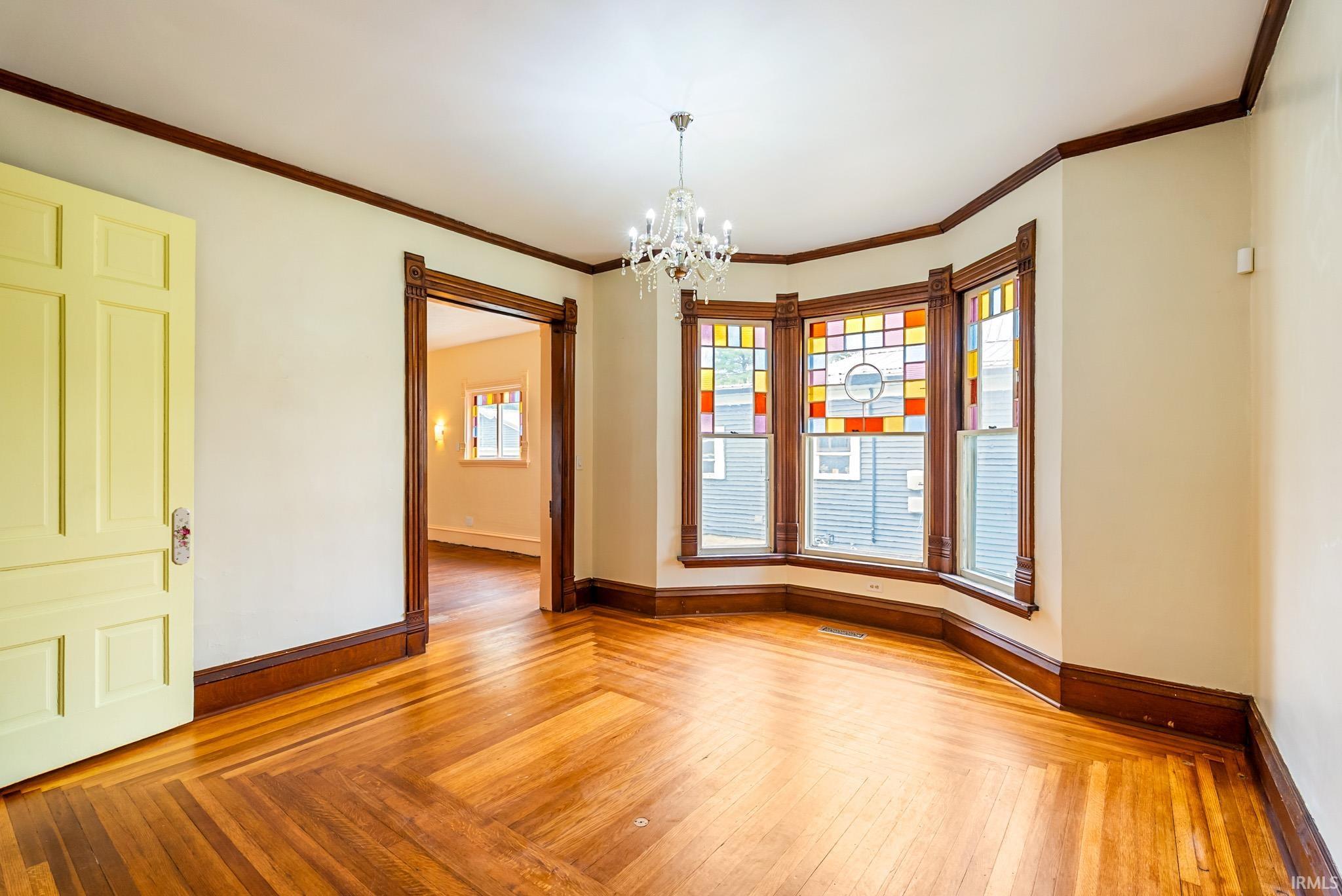 Hardwood flooring with a herringbone pattern, bay windows featuring stained glass, intricate dark wood trim, a chandelier, and a paneled interior door