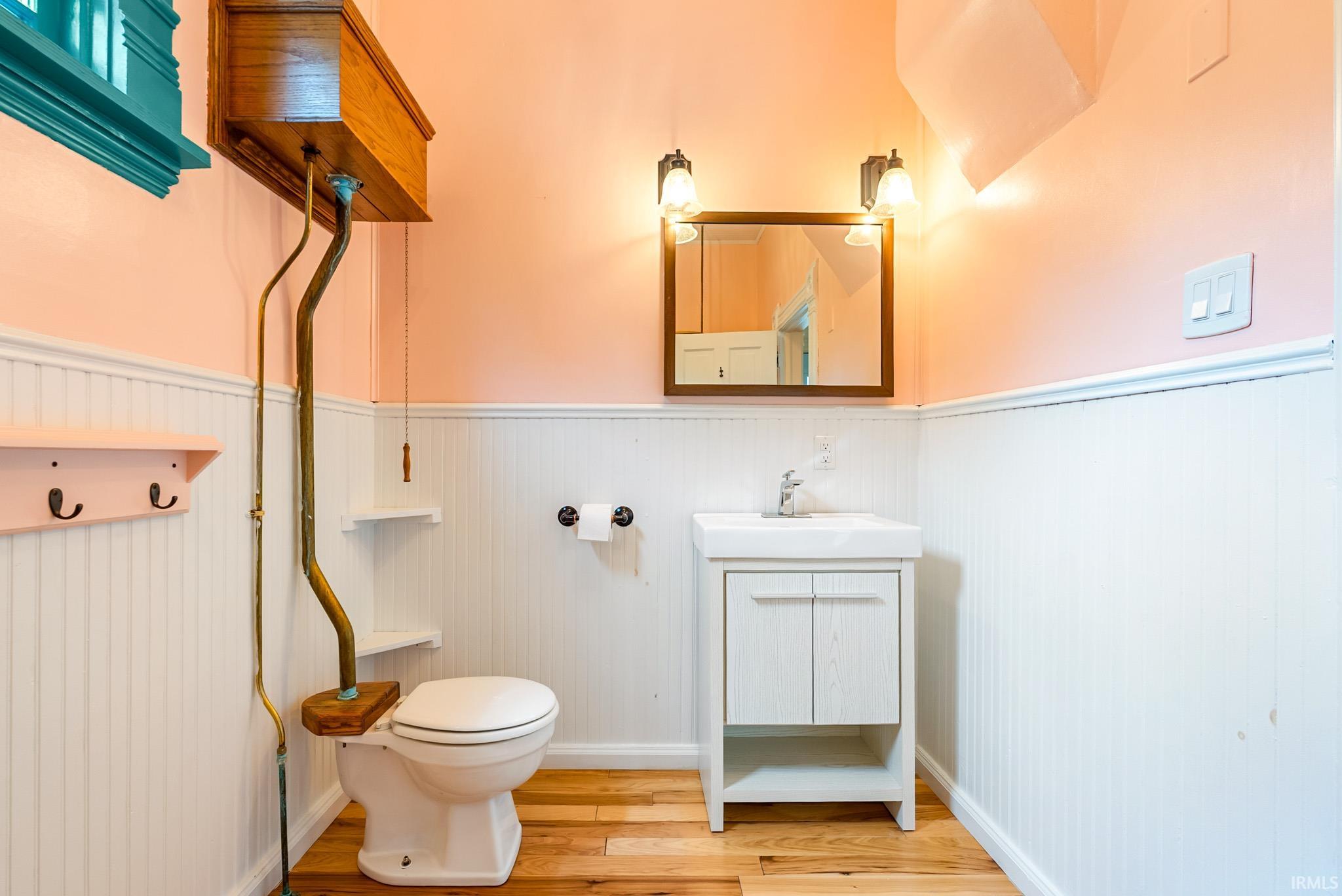 Bathroom featuring wood-finish flooring, white beadboard wainscoting, and a high-tank toilet with visible brass plumbing