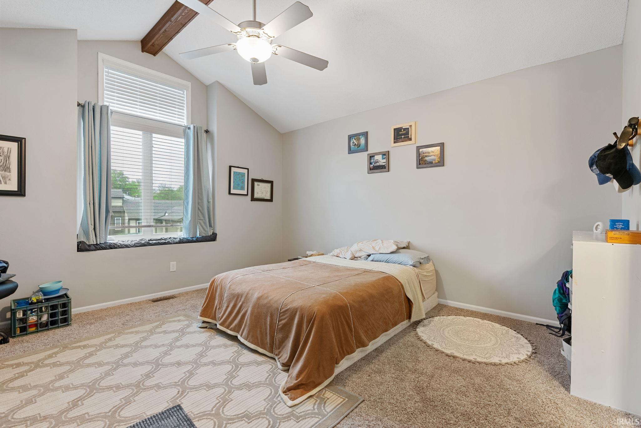 Carpeted room featuring a vaulted ceiling with exposed wooden beam, overhead fan light fixture, and a large window with horizontal blinds and drapery