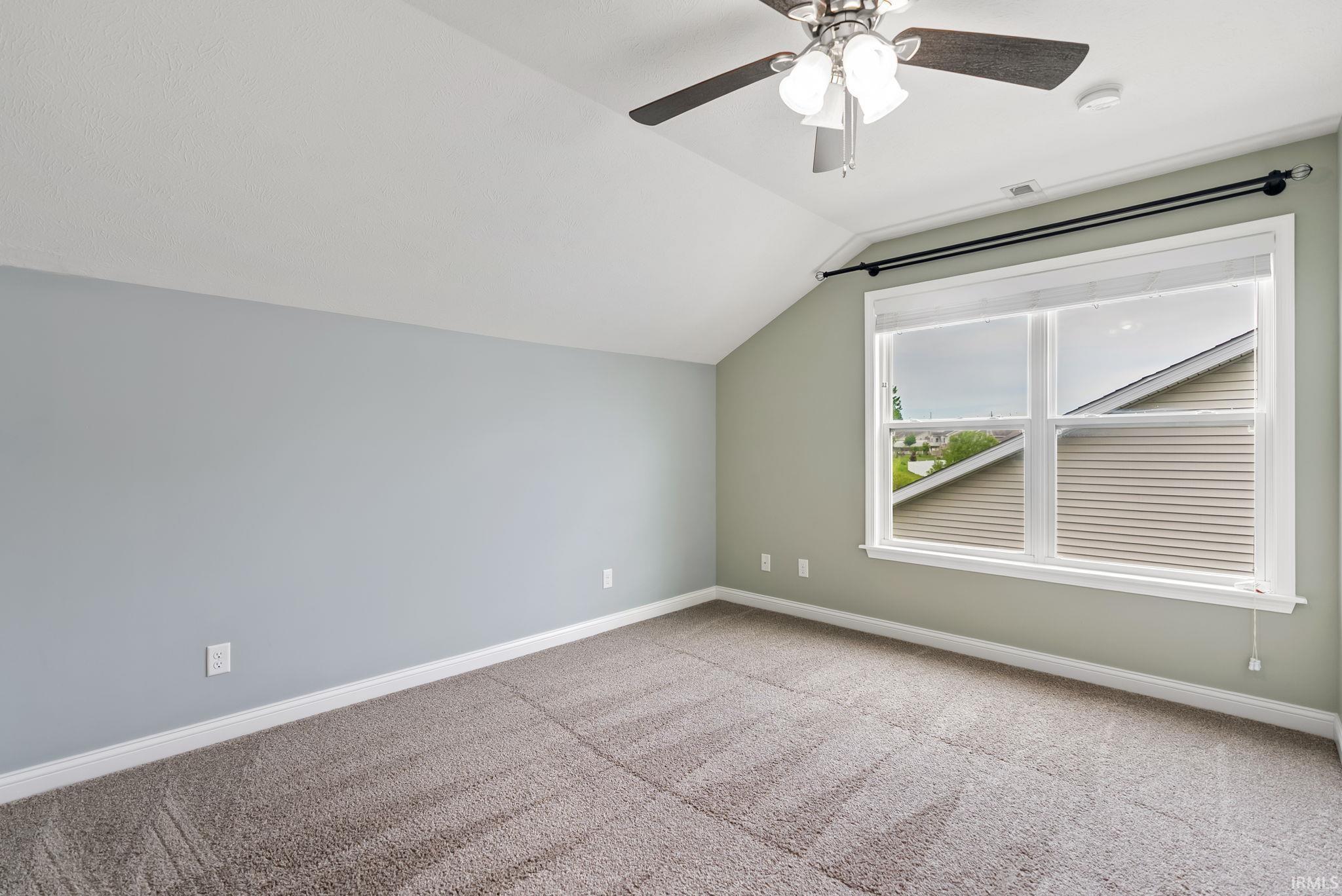 Carpeted room with a vaulted ceiling, a ceiling fan with integrated lighting, and a large window with white blinds