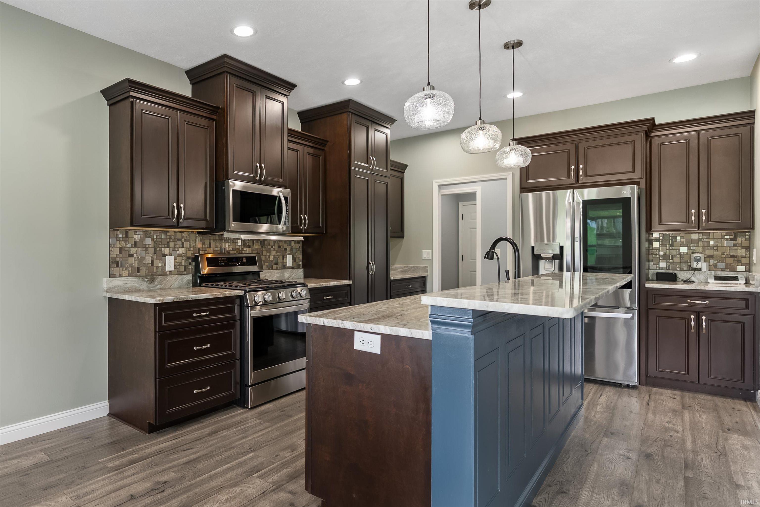 Modern kitchen featuring dark wood cabinetry, stainless steel appliances, a center island with a light-toned countertop, a mosaic tile backsplash, and wood-finish flooring