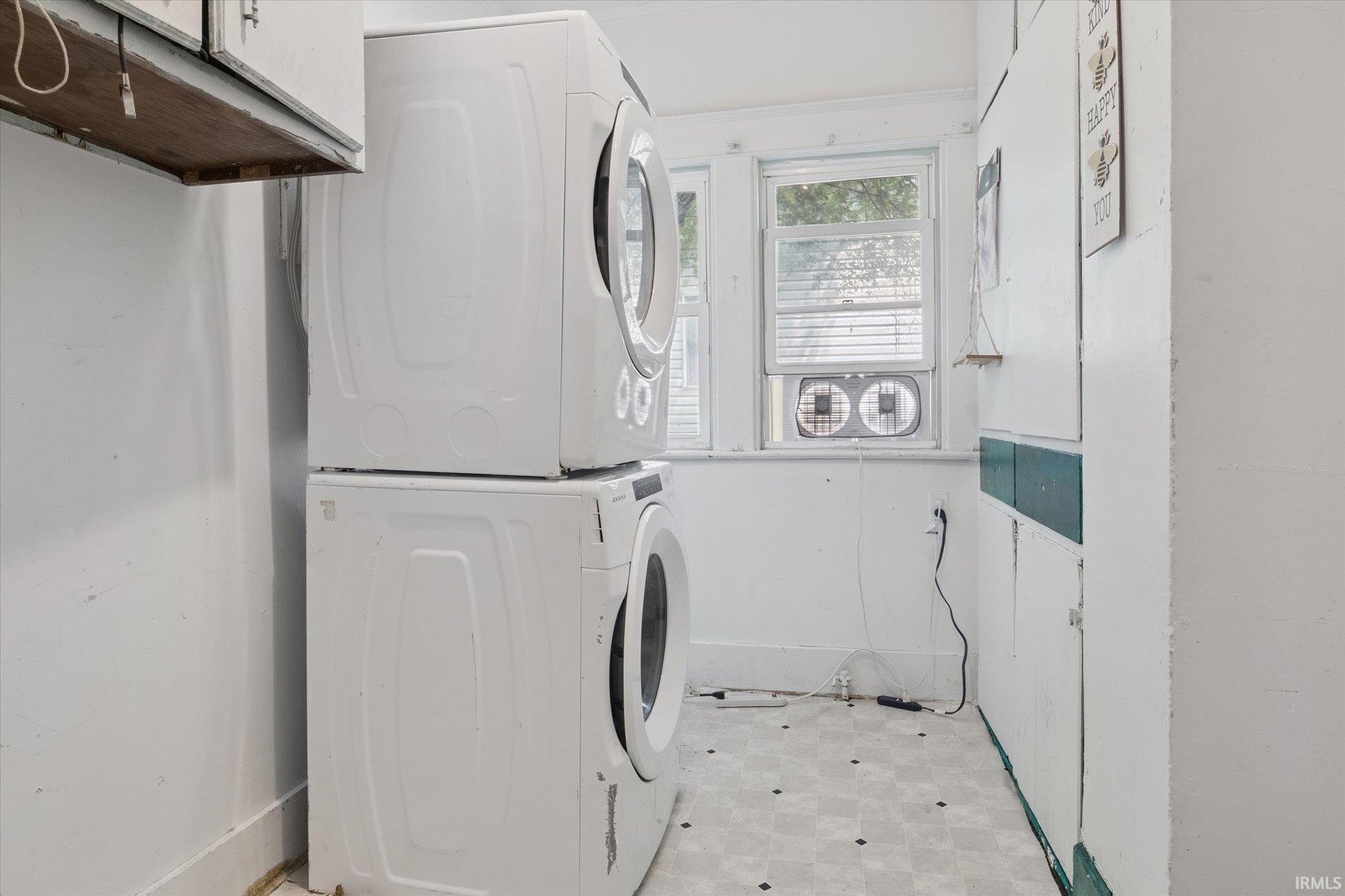 Utility space featuring stacked appliances, a window with horizontal blinds, and a patterned floor