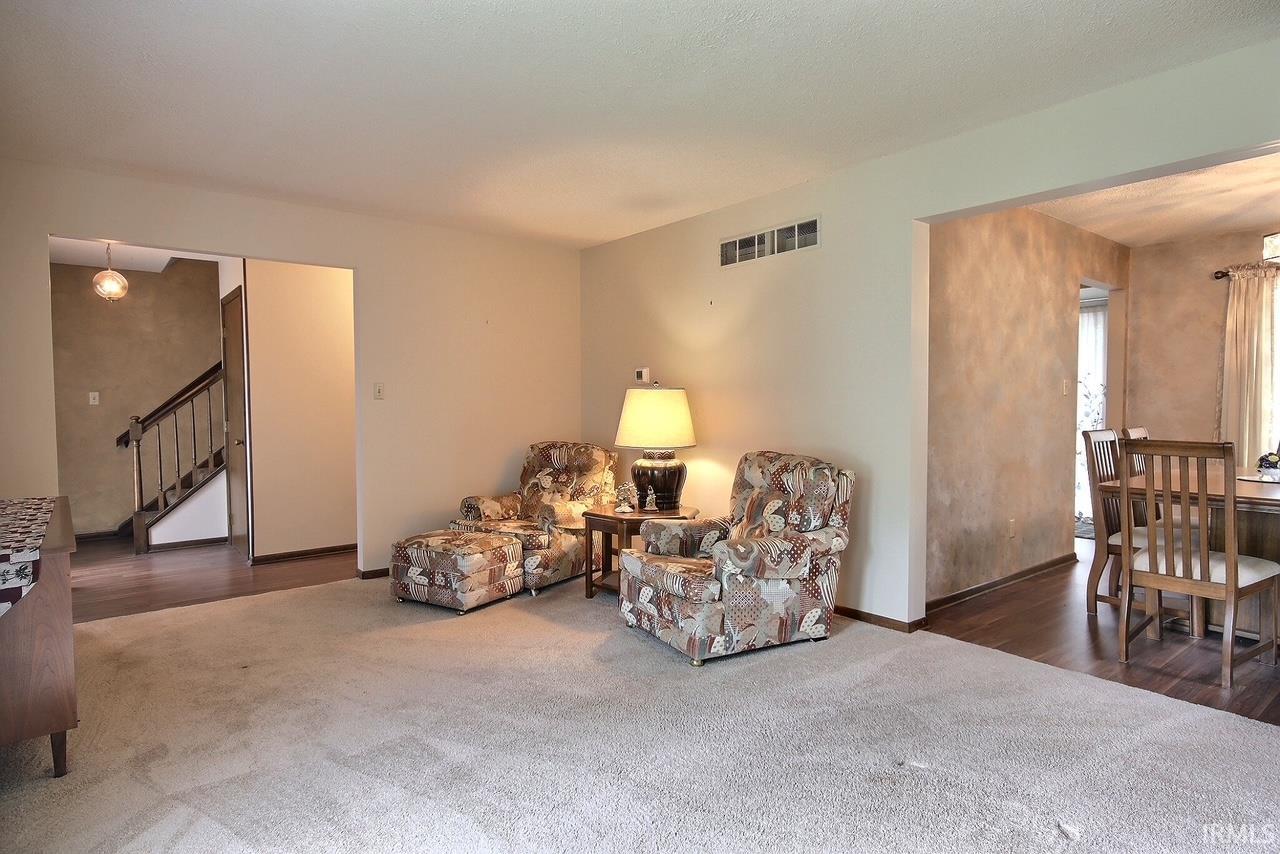Carpeted living area with light-toned walls and an open doorway to a dining space featuring wood-finish flooring