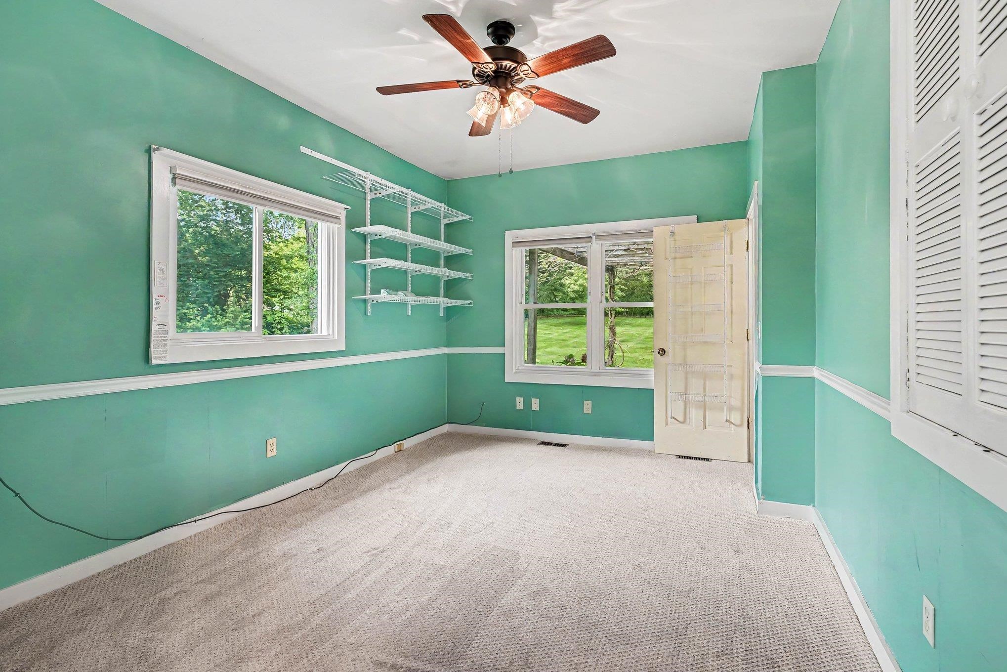 Room featuring a ceiling fan with integrated lighting, two windows, wall-mounted shelving, and carpet flooring