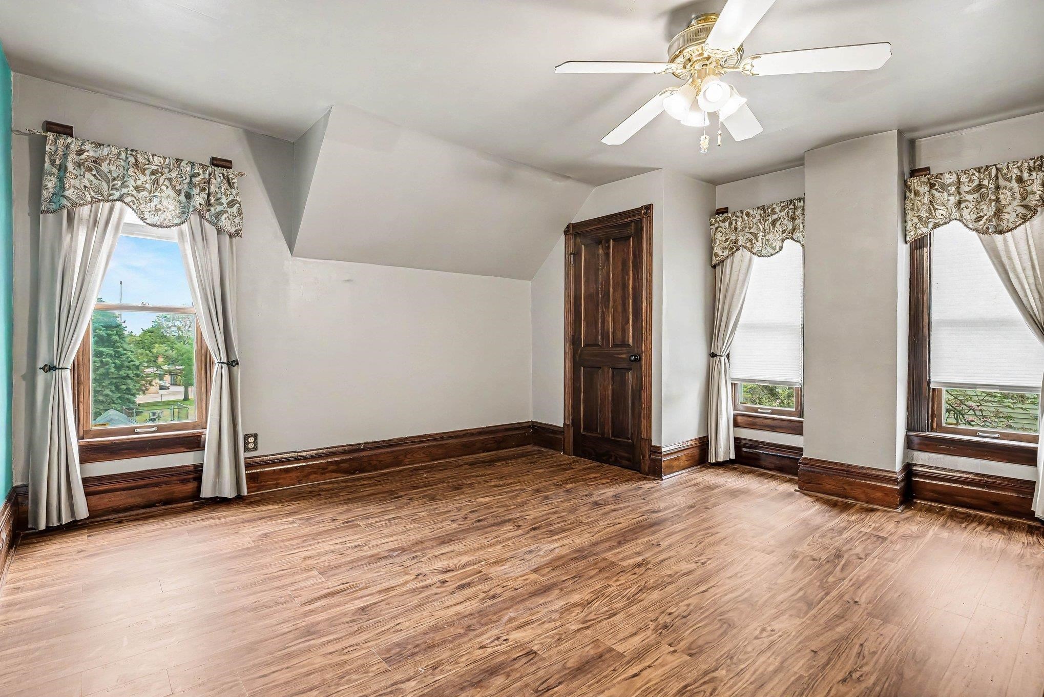 Spacious room featuring rich wood-finish flooring, a ceiling fan with integrated lighting, and multiple windows with white blinds