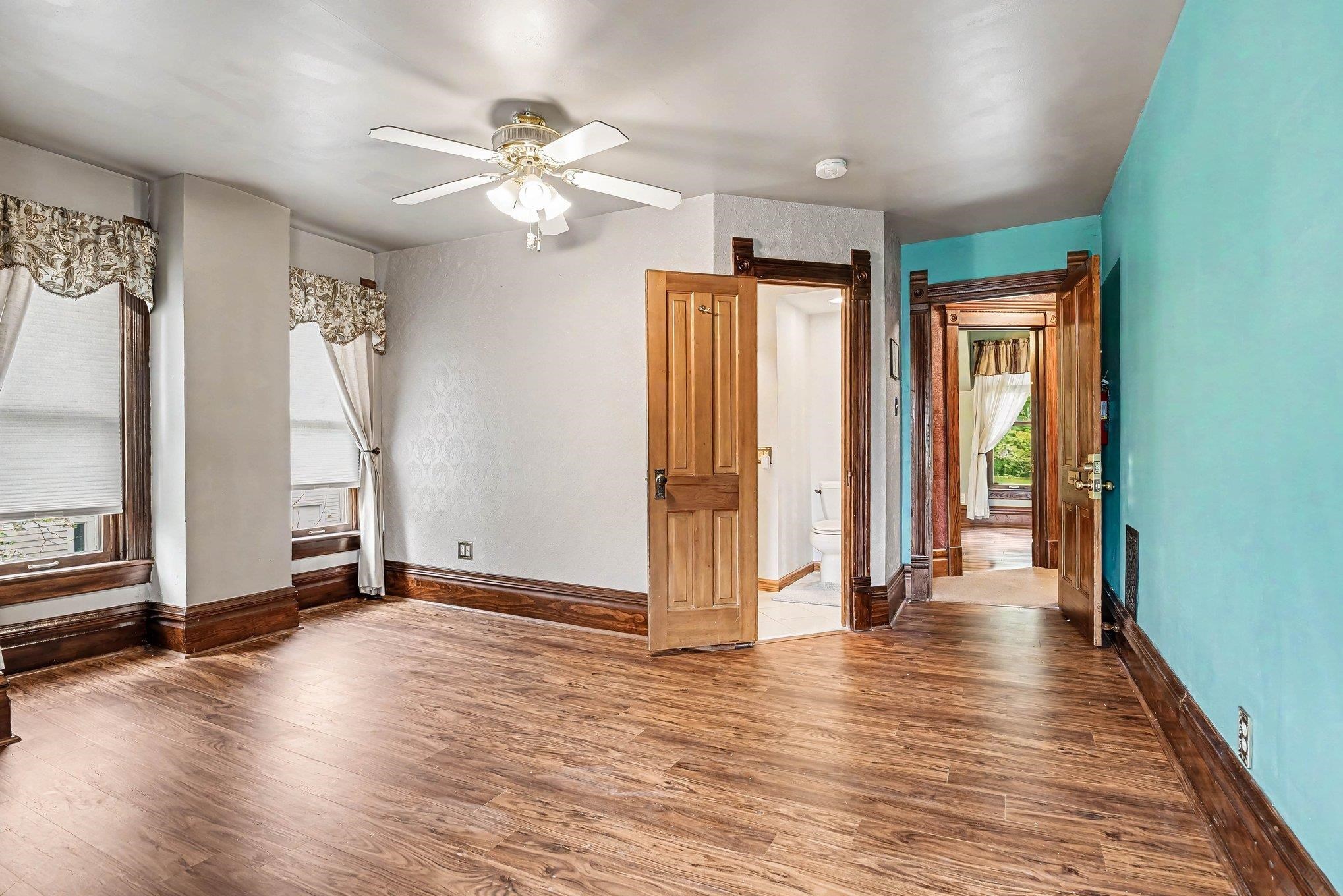 Spacious room featuring wood-finish flooring, two windows with blinds and valances, a ceiling fan with integrated lighting, and multiple paneled wood doors with dark trim