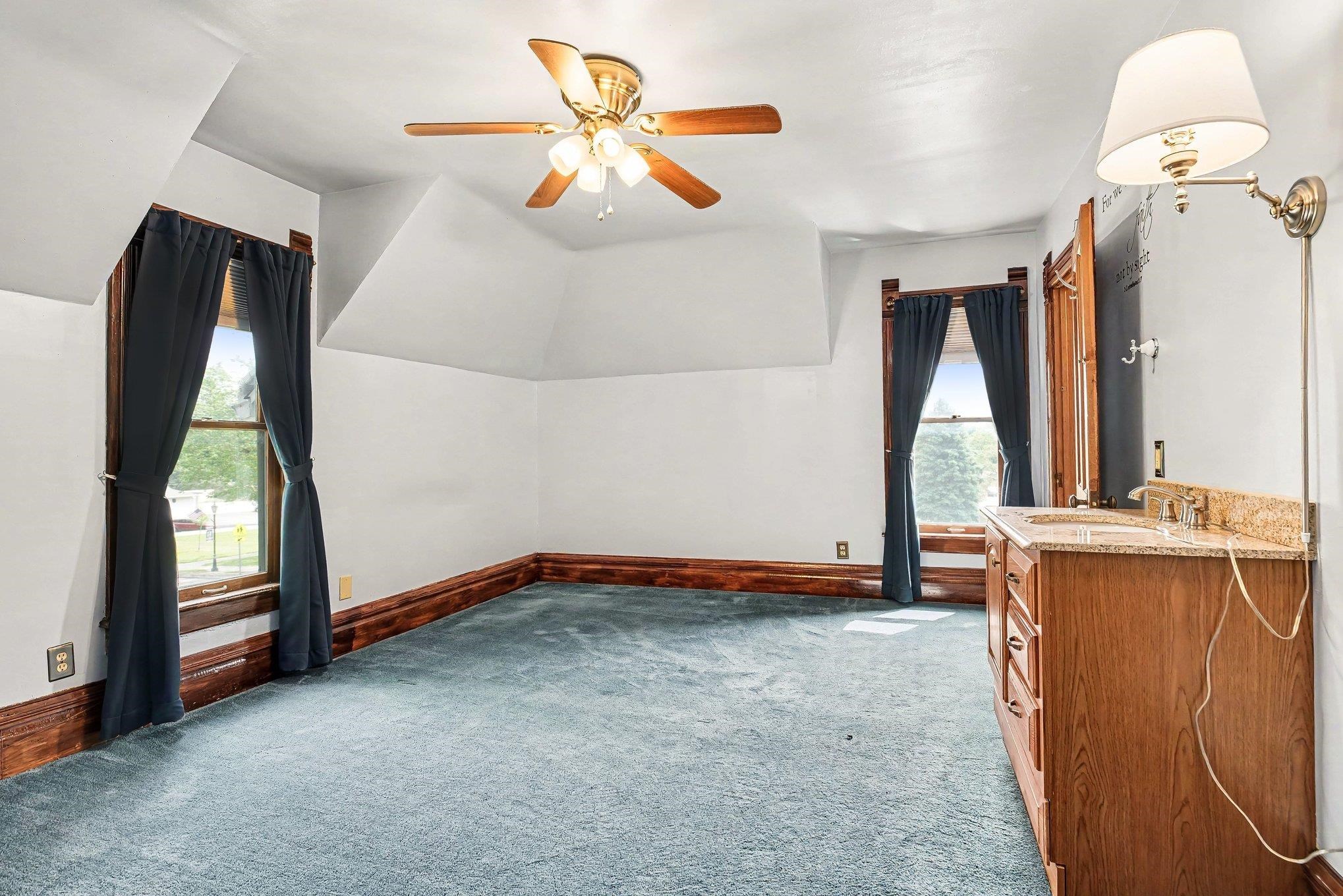 Attic space featuring sloped ceilings, two windows with wood trim, a ceiling fan, and blue carpet flooring