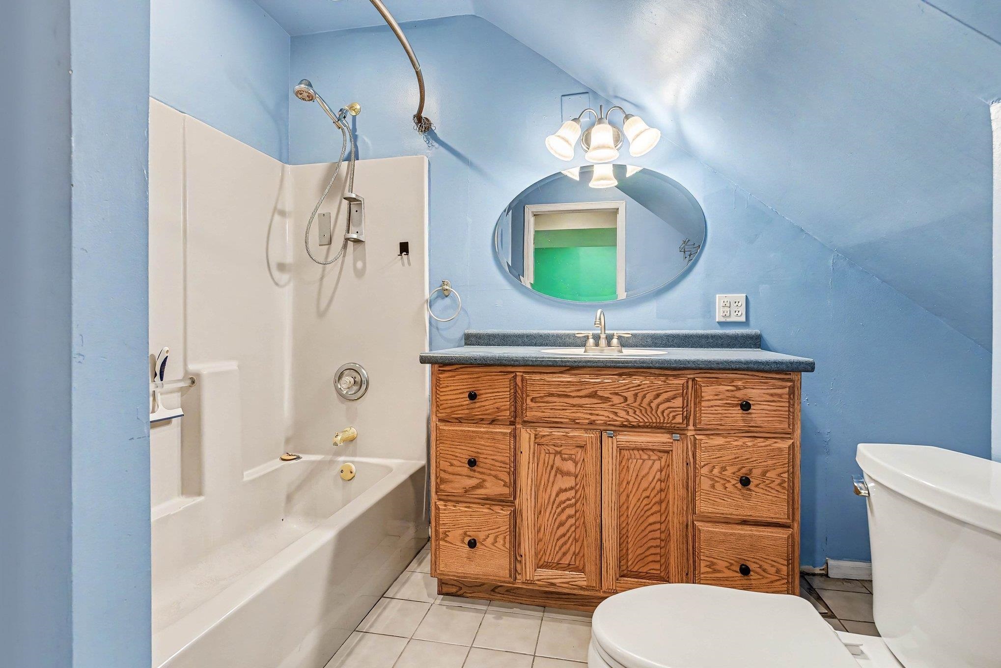 Bathroom featuring a combination tub/shower unit, a wood-finish vanity with a built-in sink, a wall-mounted oval mirror, and a white tile floor