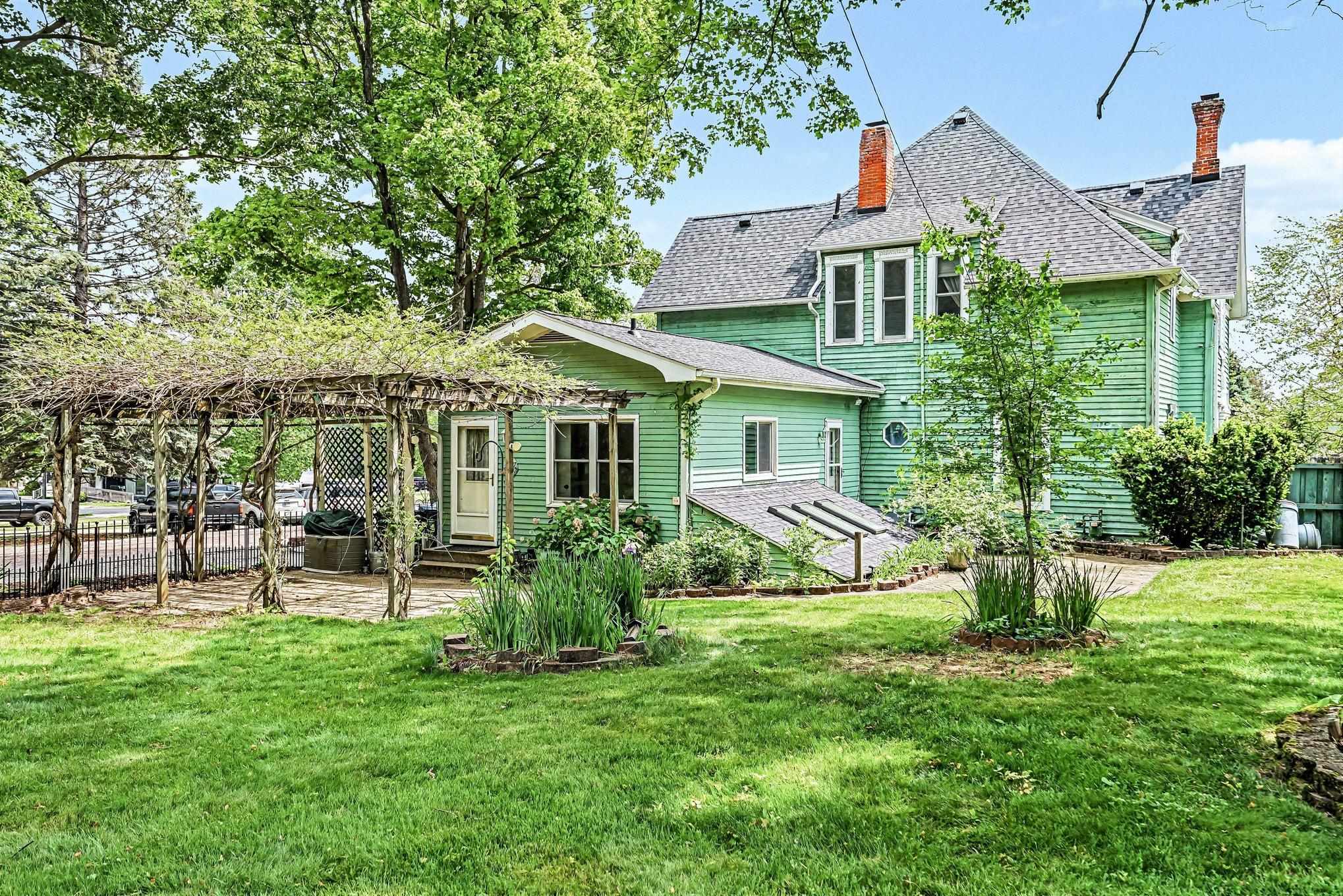Green clapboard exterior featuring a prominent pergola, stone patio, mature landscaping, and a pitched shingle roof with brick chimneys