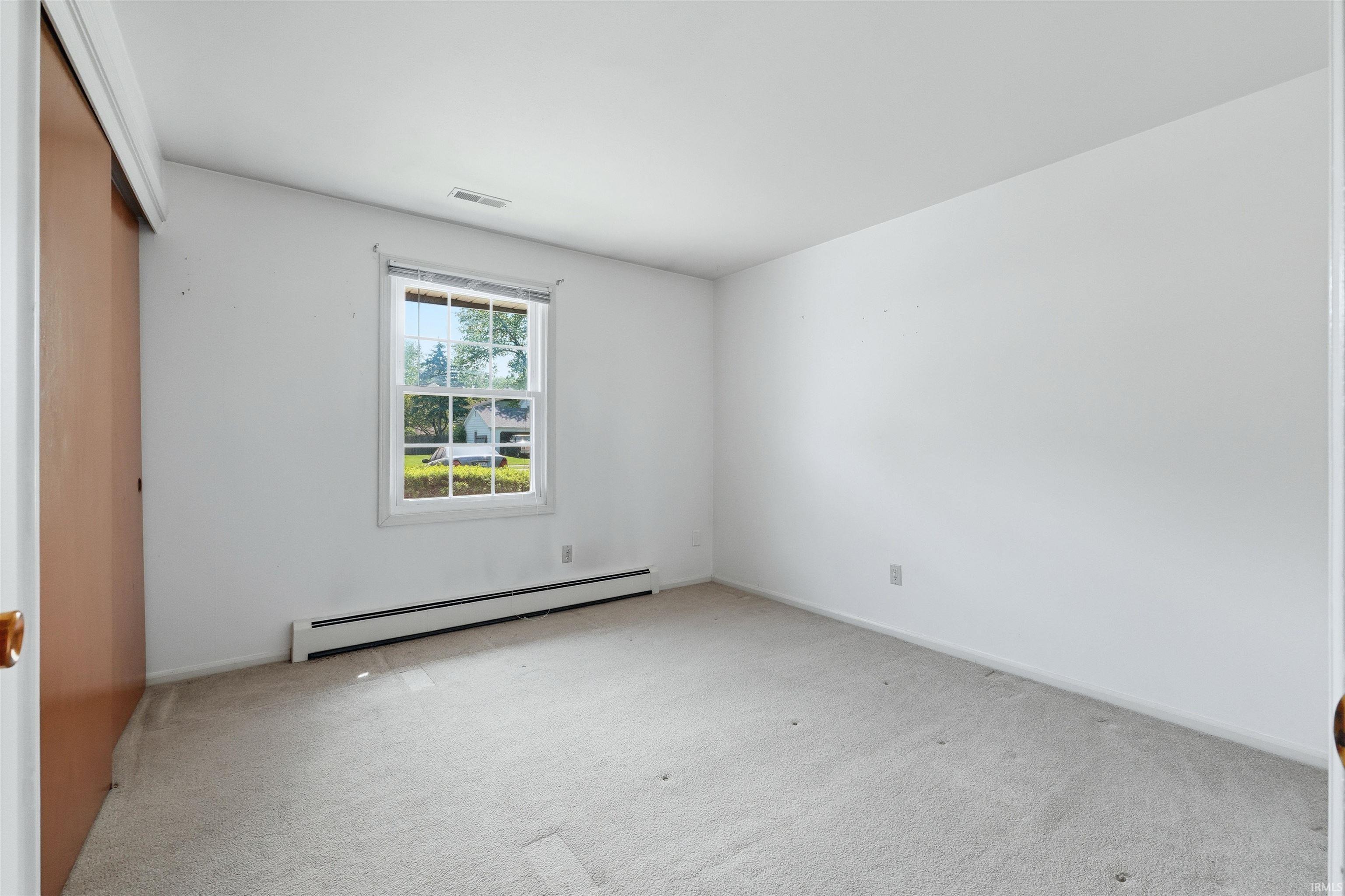 Neutral-toned carpeting paired with white walls, a single window with exterior landscaping, and a built-in closet with sliding wood-panel doors