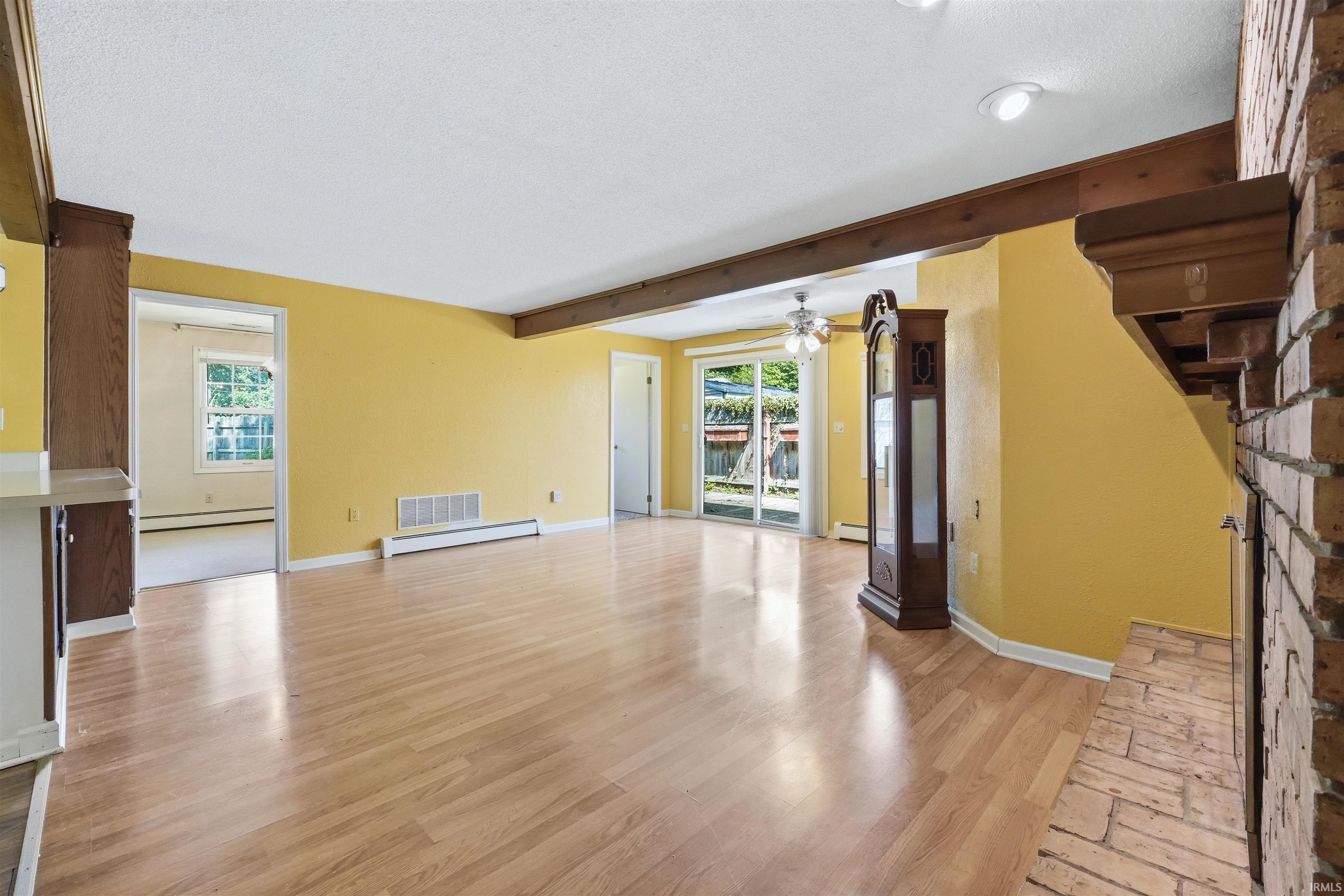Spacious room featuring wood-finish flooring, exposed ceiling beams, and a brick fireplace with a substantial mantel