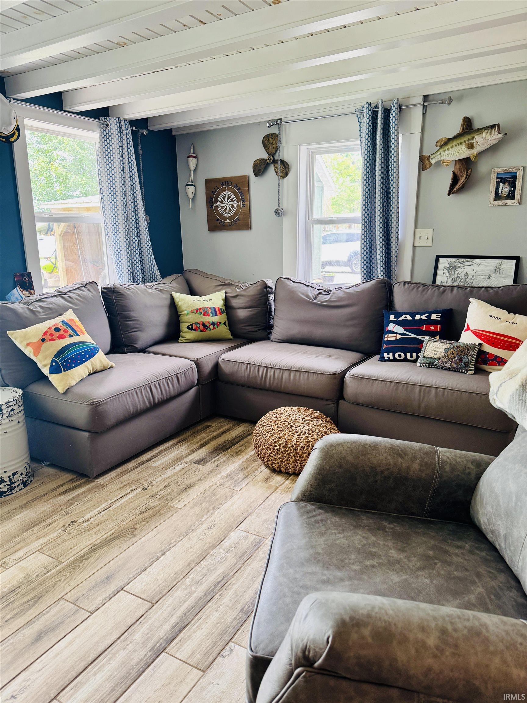 Living area featuring wood-finish flooring, exposed white ceiling beams, and dual windows allowing natural light