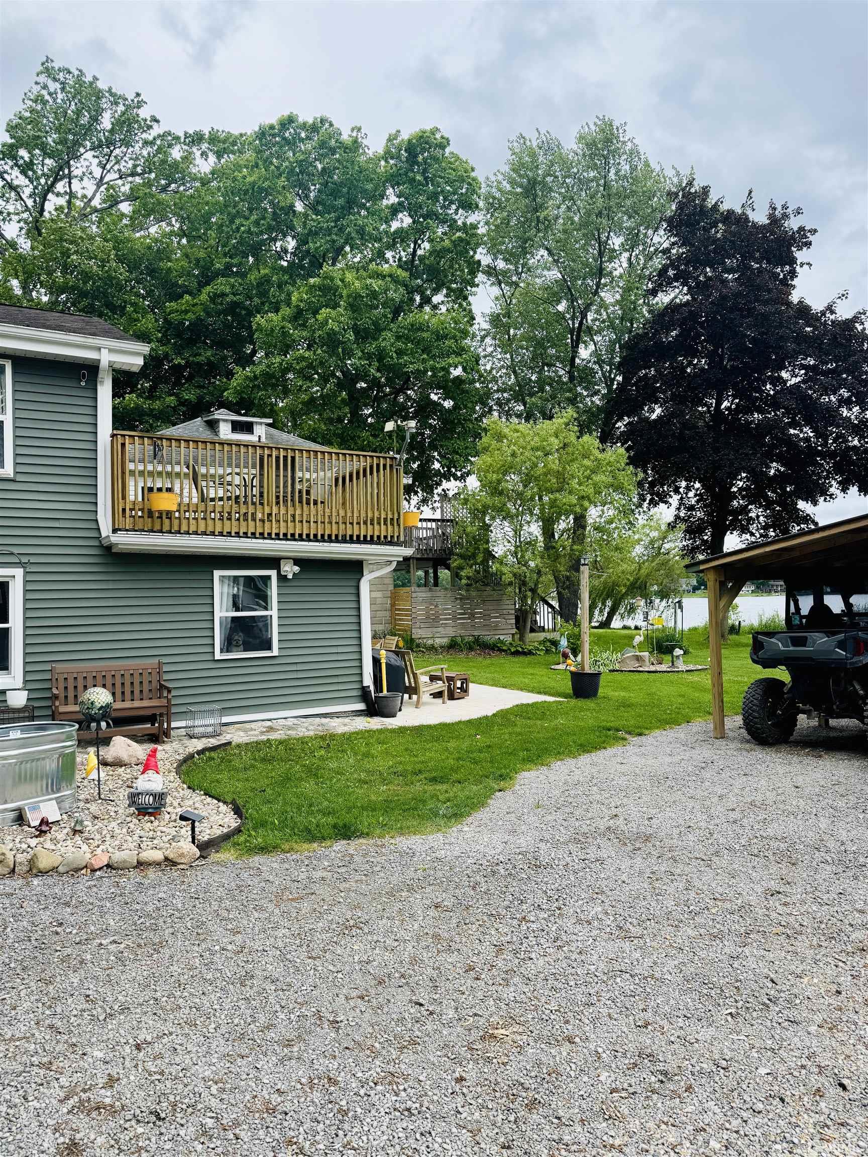 Exterior featuring a second-story wood deck, green siding, a gravel driveway, a grassy lawn, and mature trees