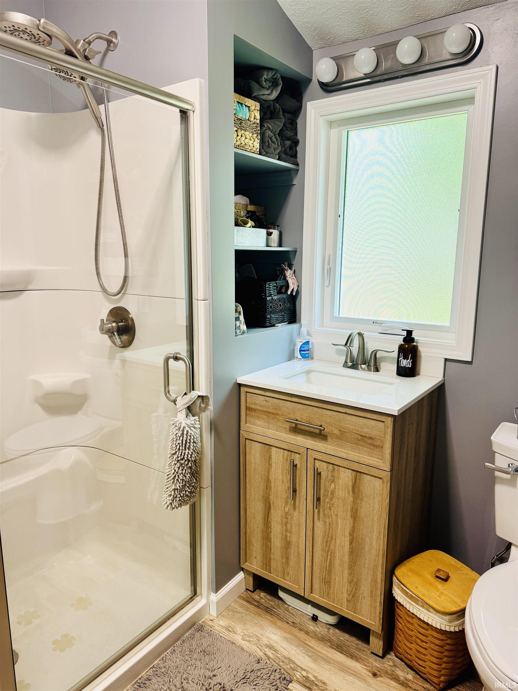 Bathroom with a walk-in shower featuring a glass enclosure, a window above the vanity, a wood-finish vanity with a white countertop, recessed shelving, and wood-finish flooring