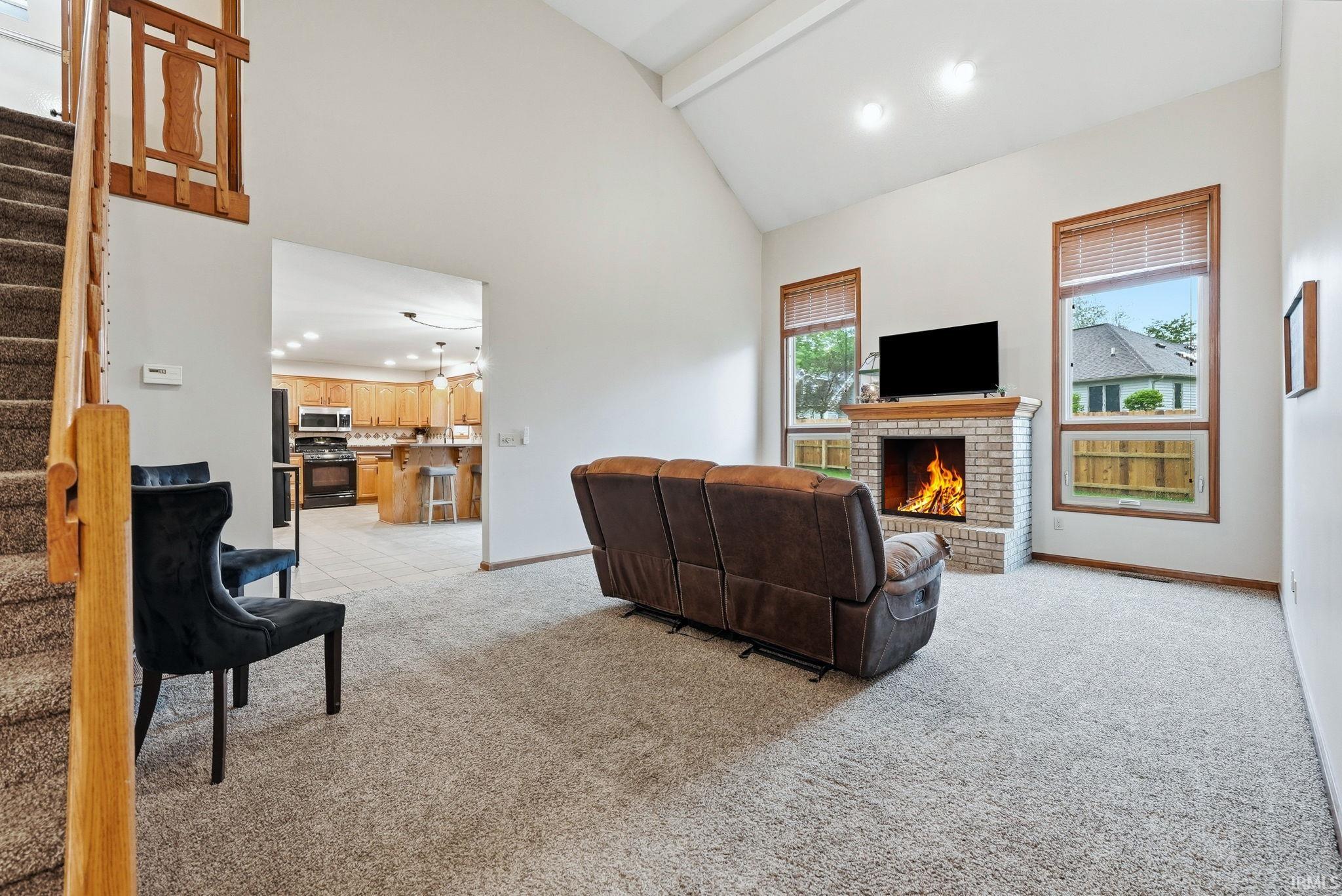 Vaulted ceiling living area featuring a brick fireplace with a wood mantle, two tall windows with wood trim, and light-colored carpeting