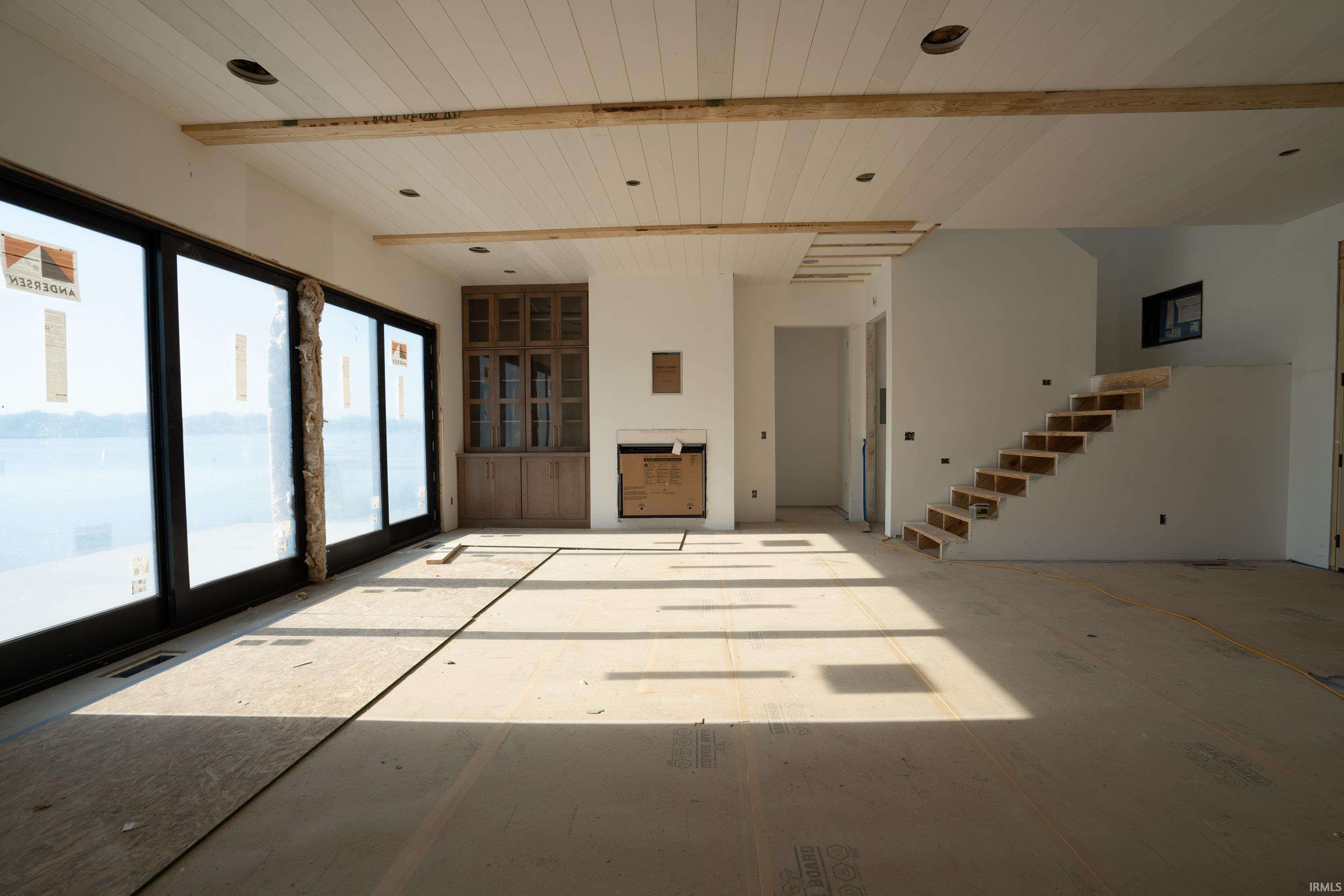 Spacious interior featuring expansive sliding glass doors, a white plank ceiling with exposed beams, recessed lighting, built-in cabinetry with glass fronts, and an open staircase