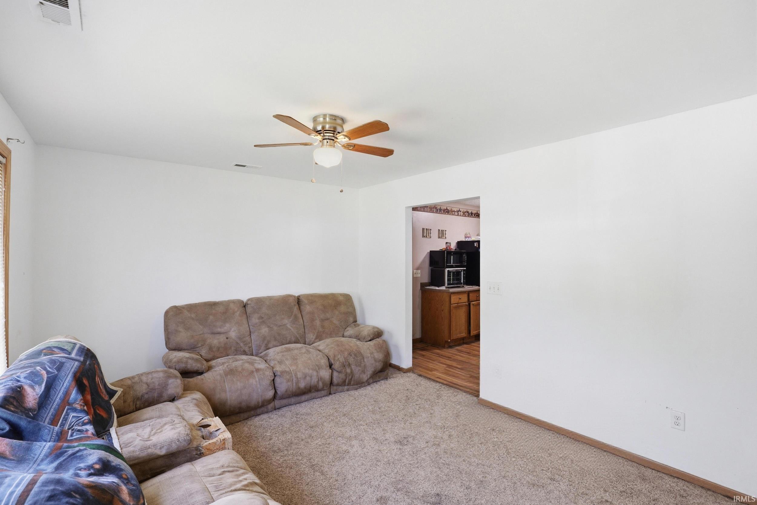 Carpeted living space featuring a ceiling fan with light fixture, white walls, and a window with wood trim