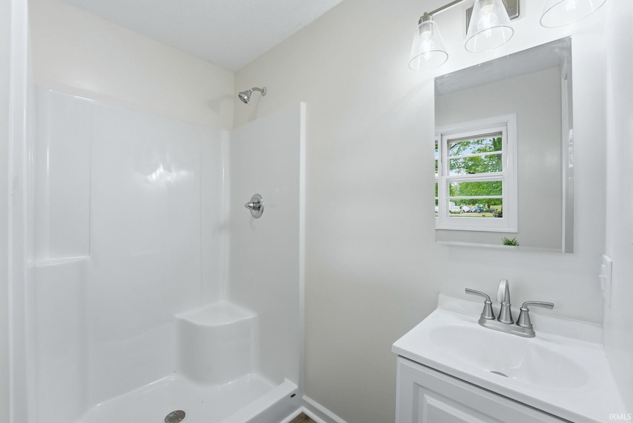 Bathroom featuring a white shower stall with built-in seating, a vanity with a white basin and chrome fixtures, a wall-mounted mirror, and a three-light vanity fixture with clear glass shades