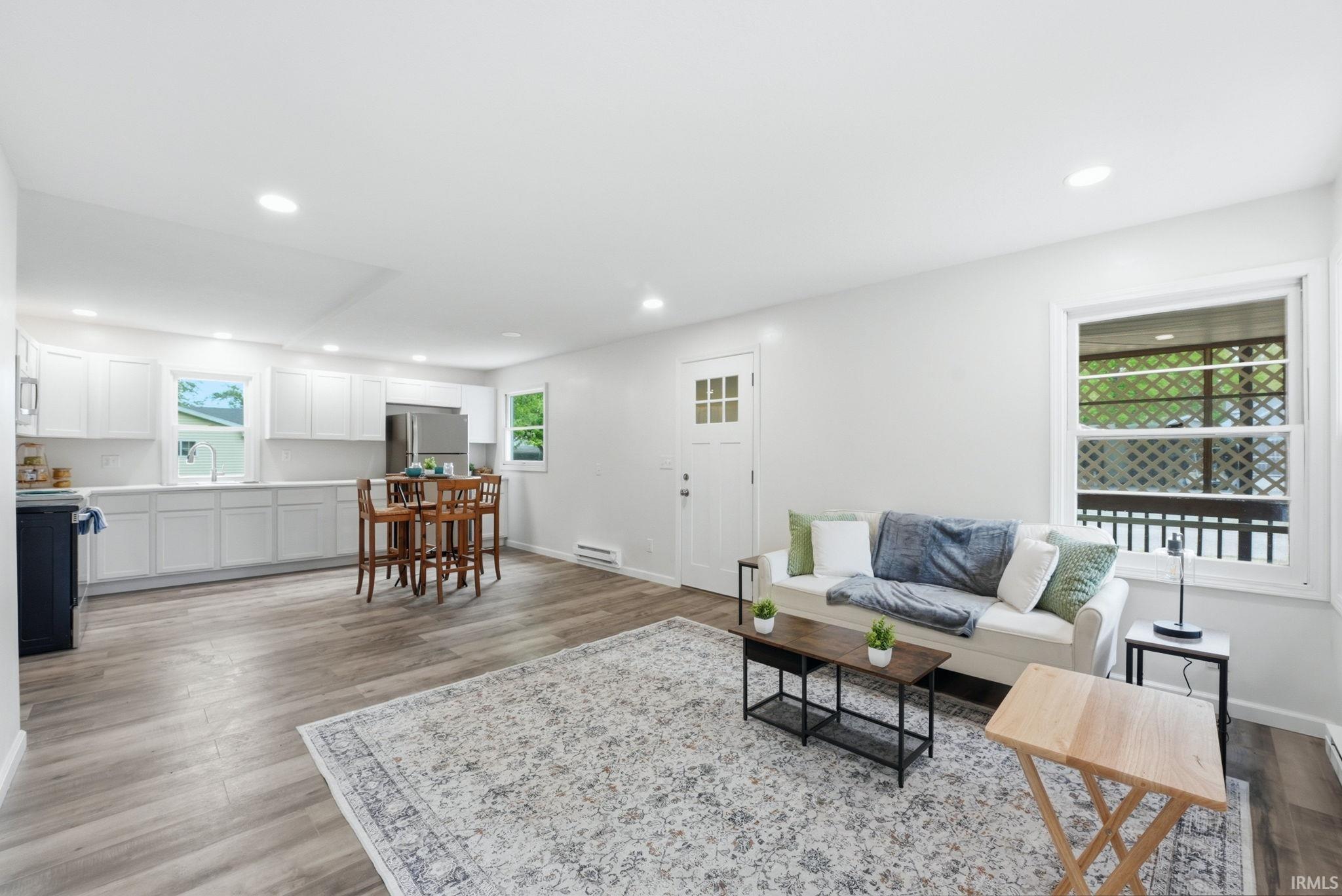 Open-concept living space featuring wood-finish flooring, recessed lighting, white shaker-style cabinetry, a built-in stainless steel refrigerator, and a kitchen sink with a window