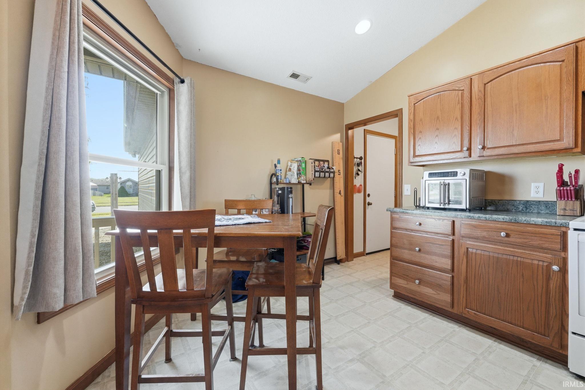 Kitchen dining area featuring a prominent window with wood trim, wood-finish cabinetry, speckled countertops, and vaulted ceilings
