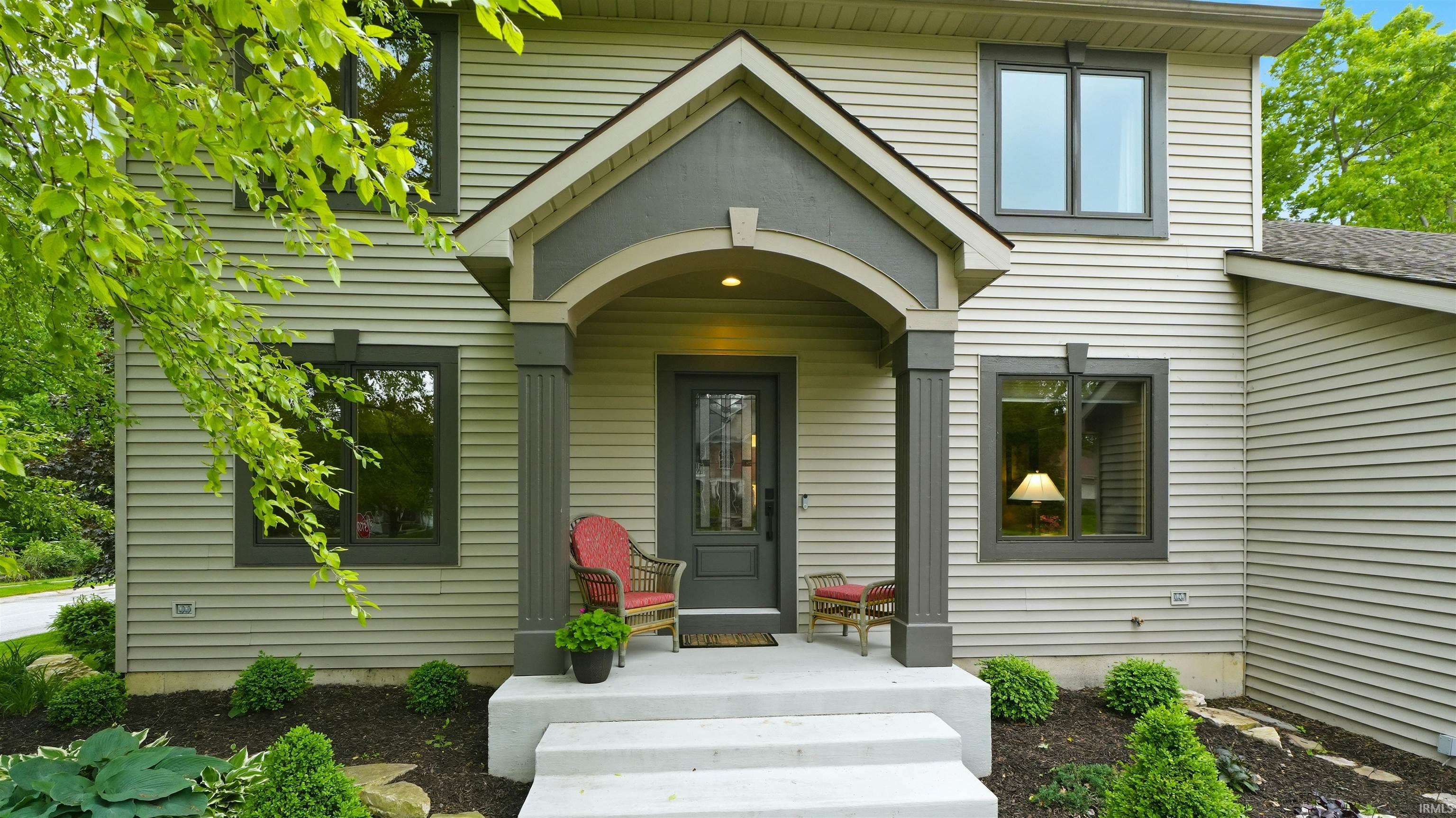 Exterior facade featuring horizontal siding, dark-trimmed windows, a prominent covered entryway with decorative columns, and mature landscaping