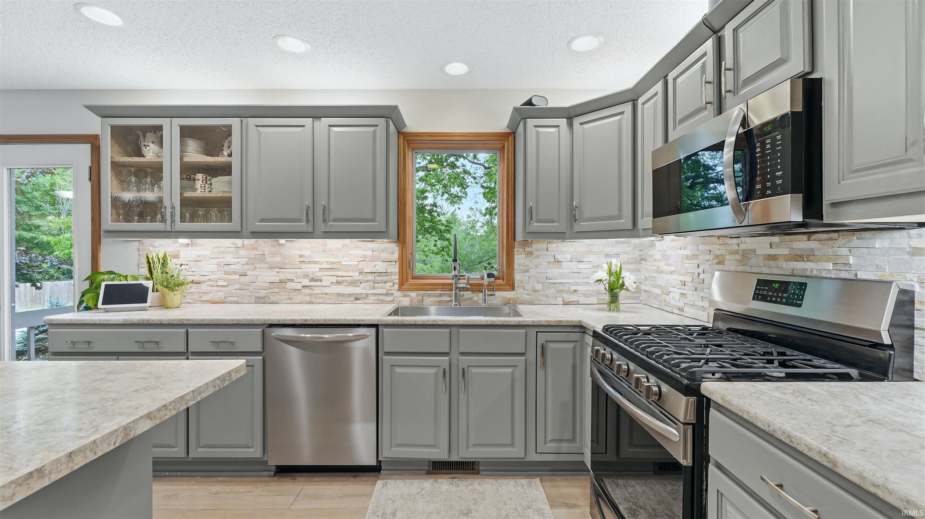 Kitchen featuring gray cabinetry with brushed nickel hardware, light-toned stone-finish countertops, stainless steel appliances, a stacked stone backsplash, and recessed lighting