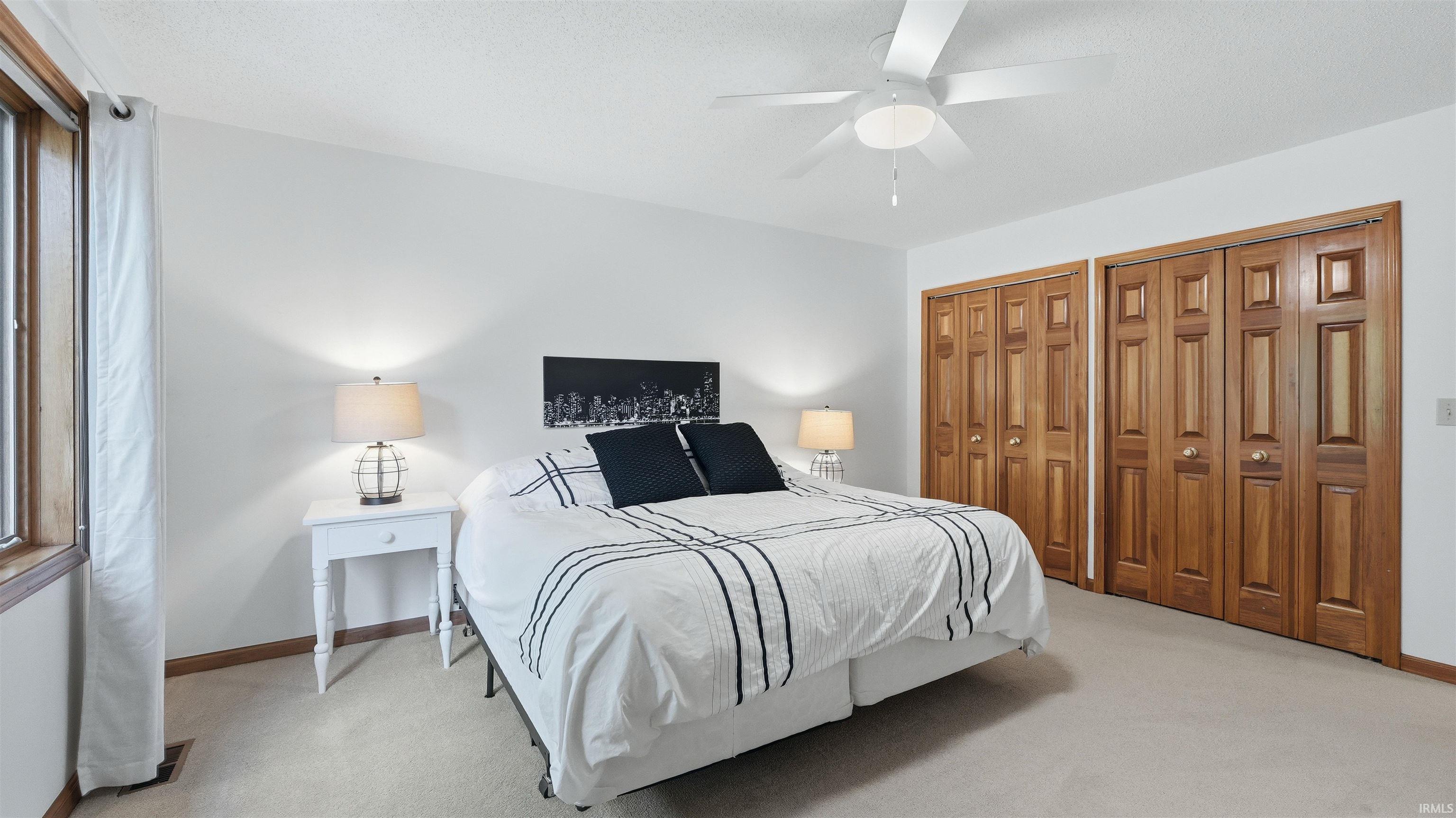 Bright bedroom featuring a ceiling fan, two paneled wood bifold closet doors, light neutral carpeting, and a large window with wood trim
