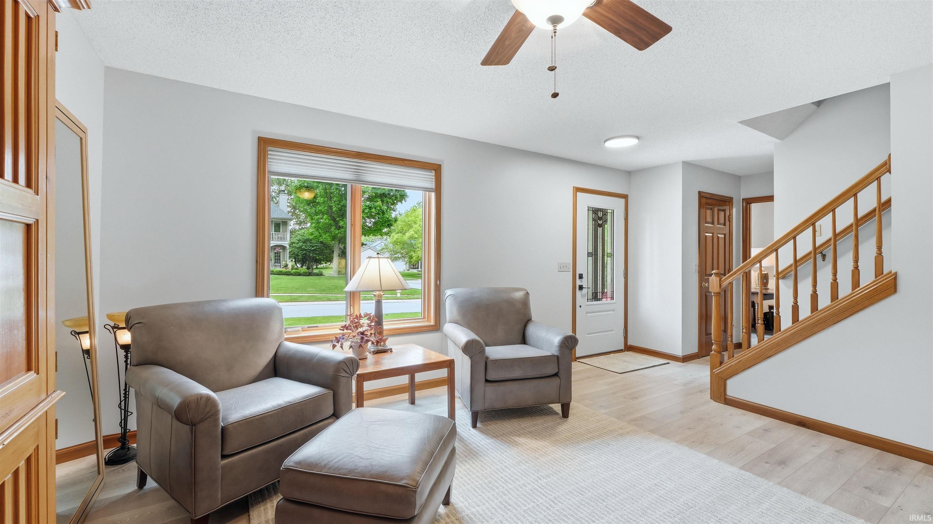 Living area featuring wood-finish flooring, a large window with wood trim, a ceiling fan with light, a white entry door with glass inserts, and a staircase with wood railings
