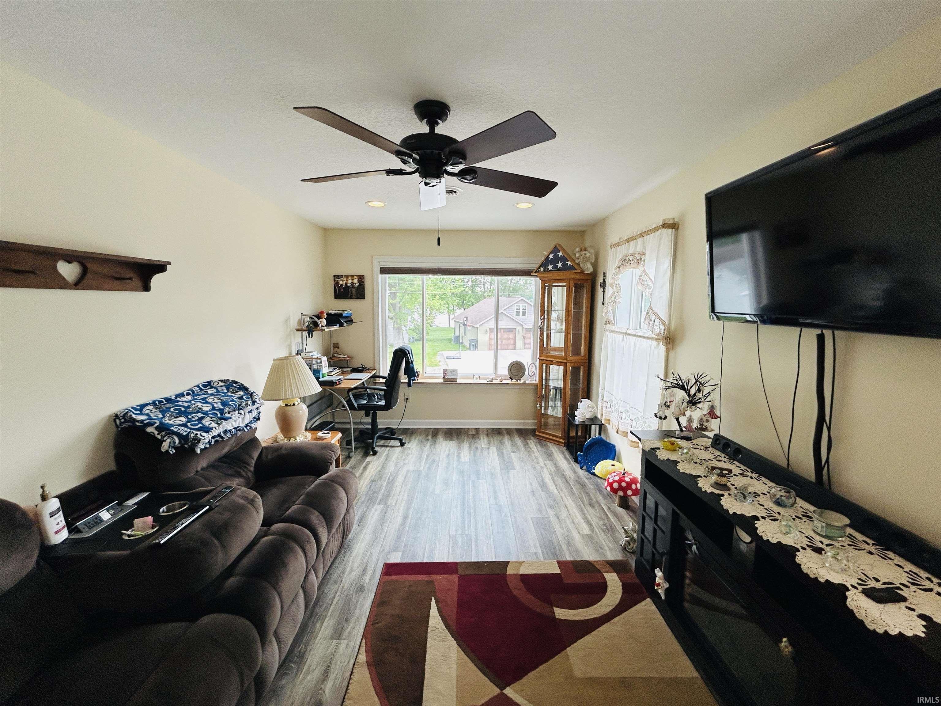 Spacious living area featuring wood-finish flooring, a ceiling fan with integrated lighting, and a large window providing natural light