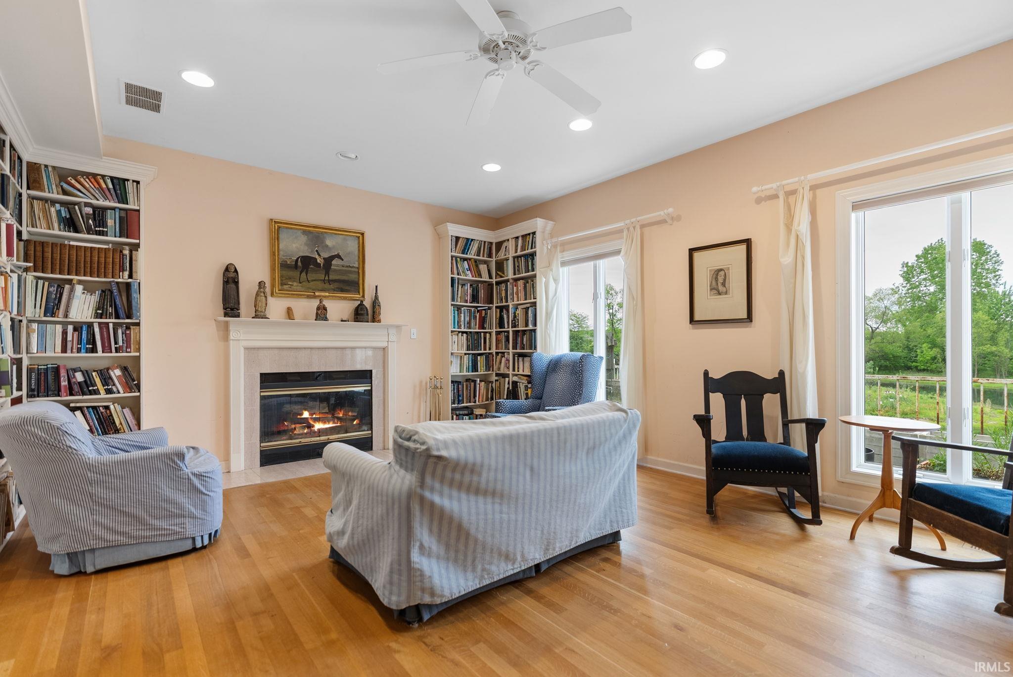 Living area featuring a fireplace with a light-toned surround and mantel, built-in bookshelves, hardwood flooring, and large windows