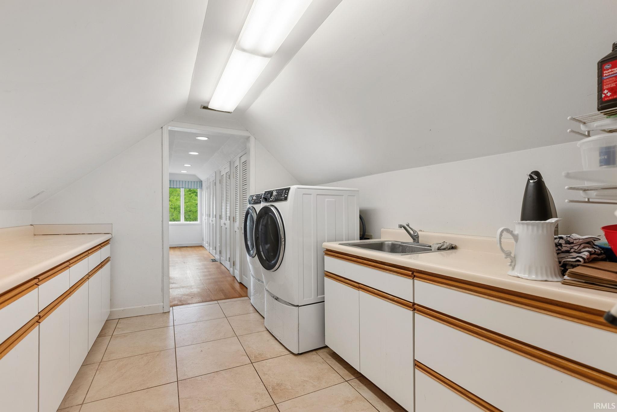 Dedicated laundry area featuring a utility sink, extensive counter space, white cabinetry with wood-finish trim, tile flooring, and sloped ceilings