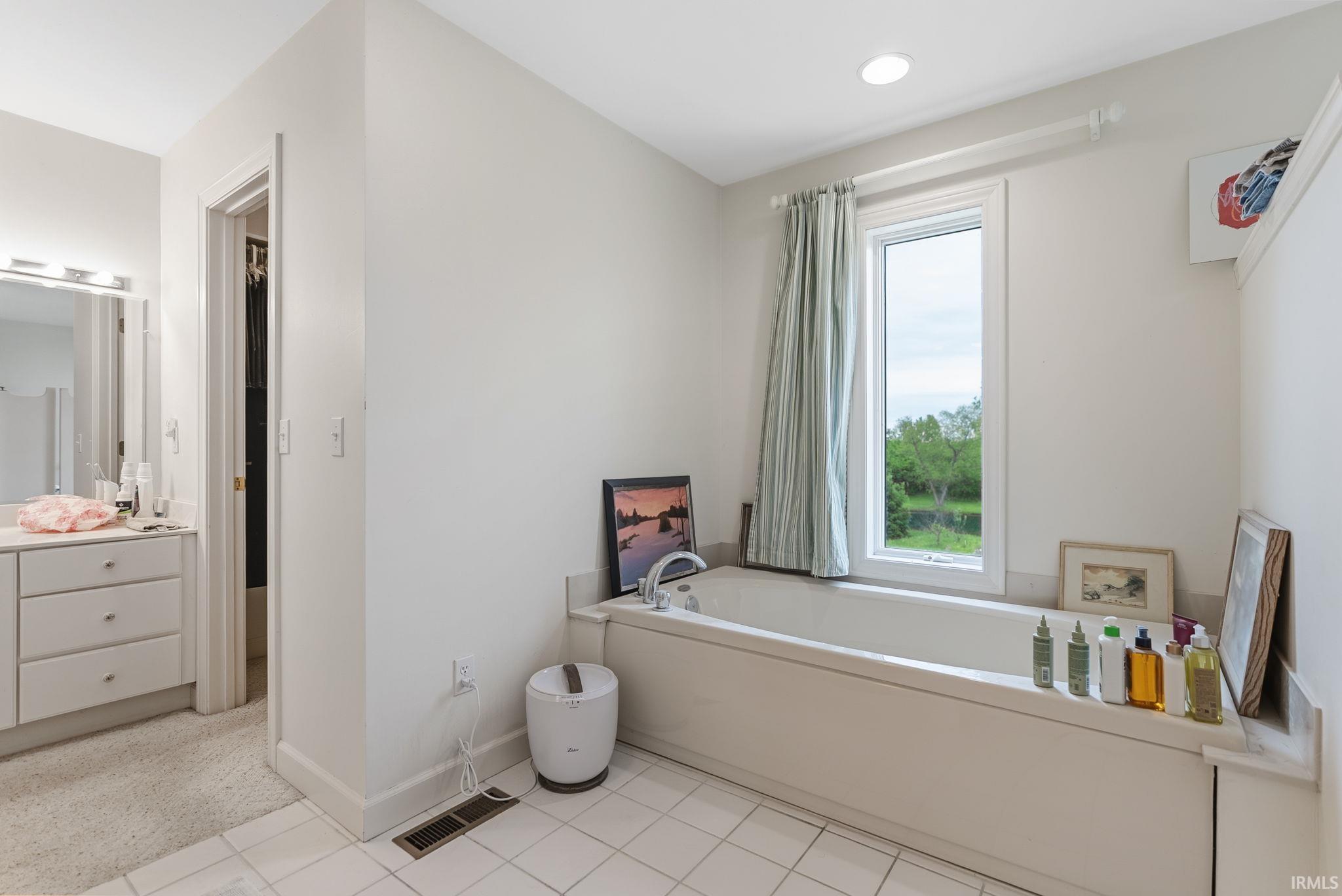Soaking tub with a window, white tile flooring, recessed lighting, and a vanity with a mirrored wall and built-in cabinetry