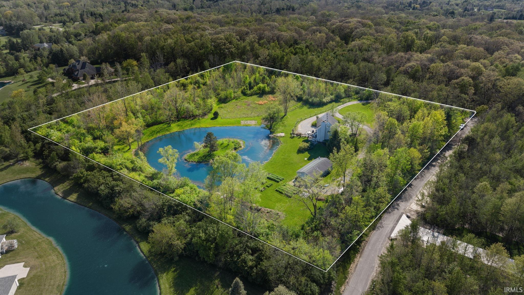 Expansive property featuring a private pond with a central island, mature trees, a winding driveway, and a greenhouse structure