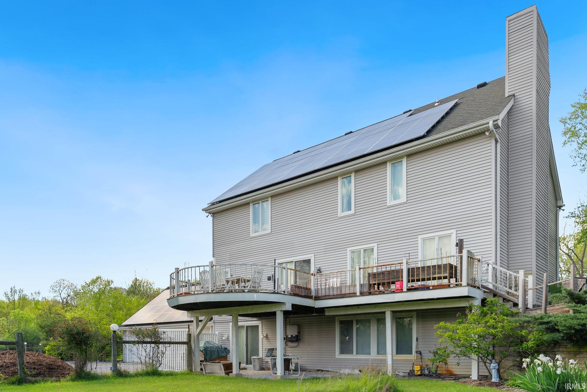 Rear exterior featuring grey horizontal siding, a multi-level deck with wood railing, solar panels on the roof, a large exterior chimney, and a lower-level patio with sliding glass doors