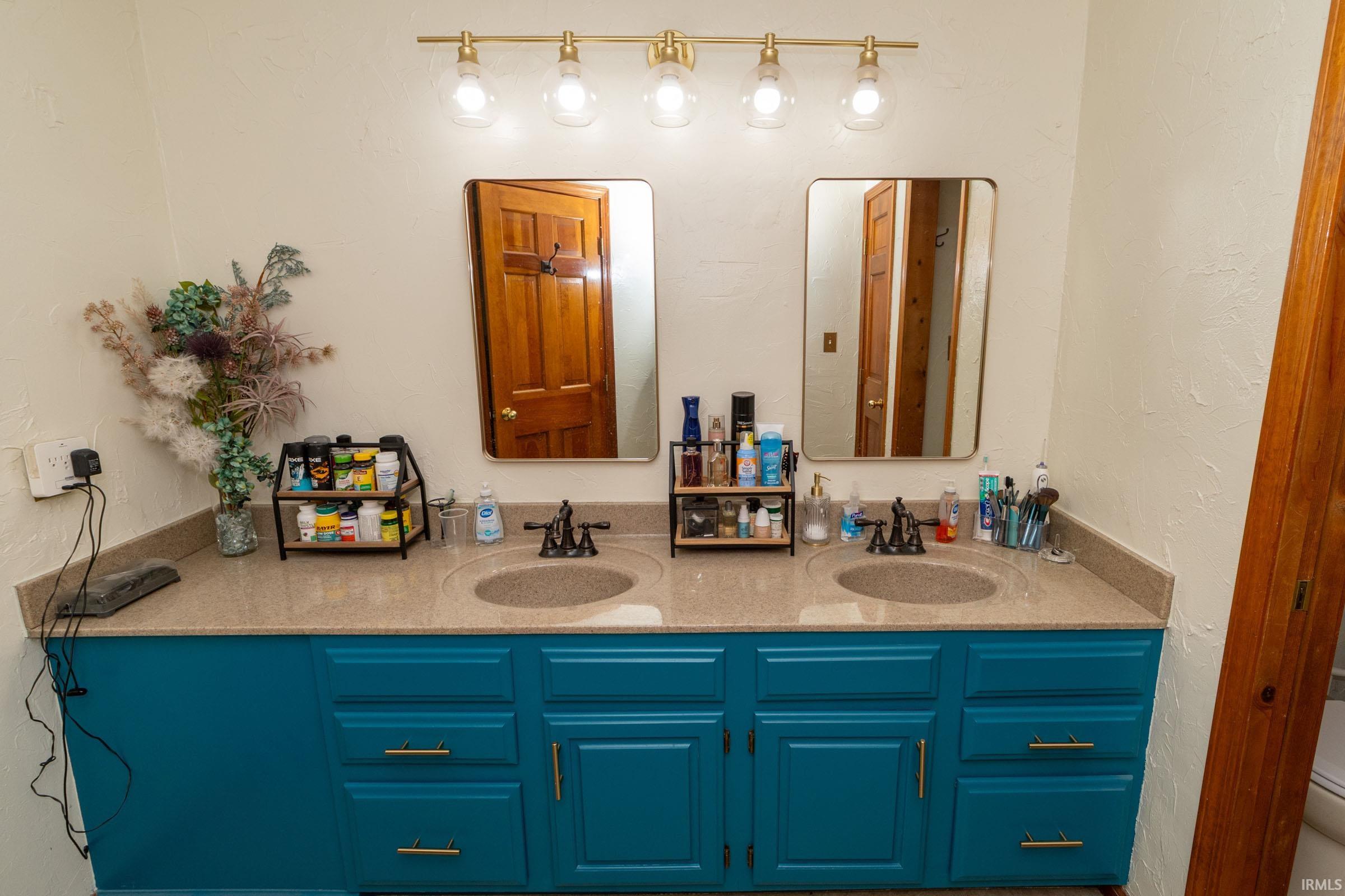Dual sink vanity featuring teal cabinetry with gold-toned hardware, a light-toned countertop, and matching rectangular mirrors