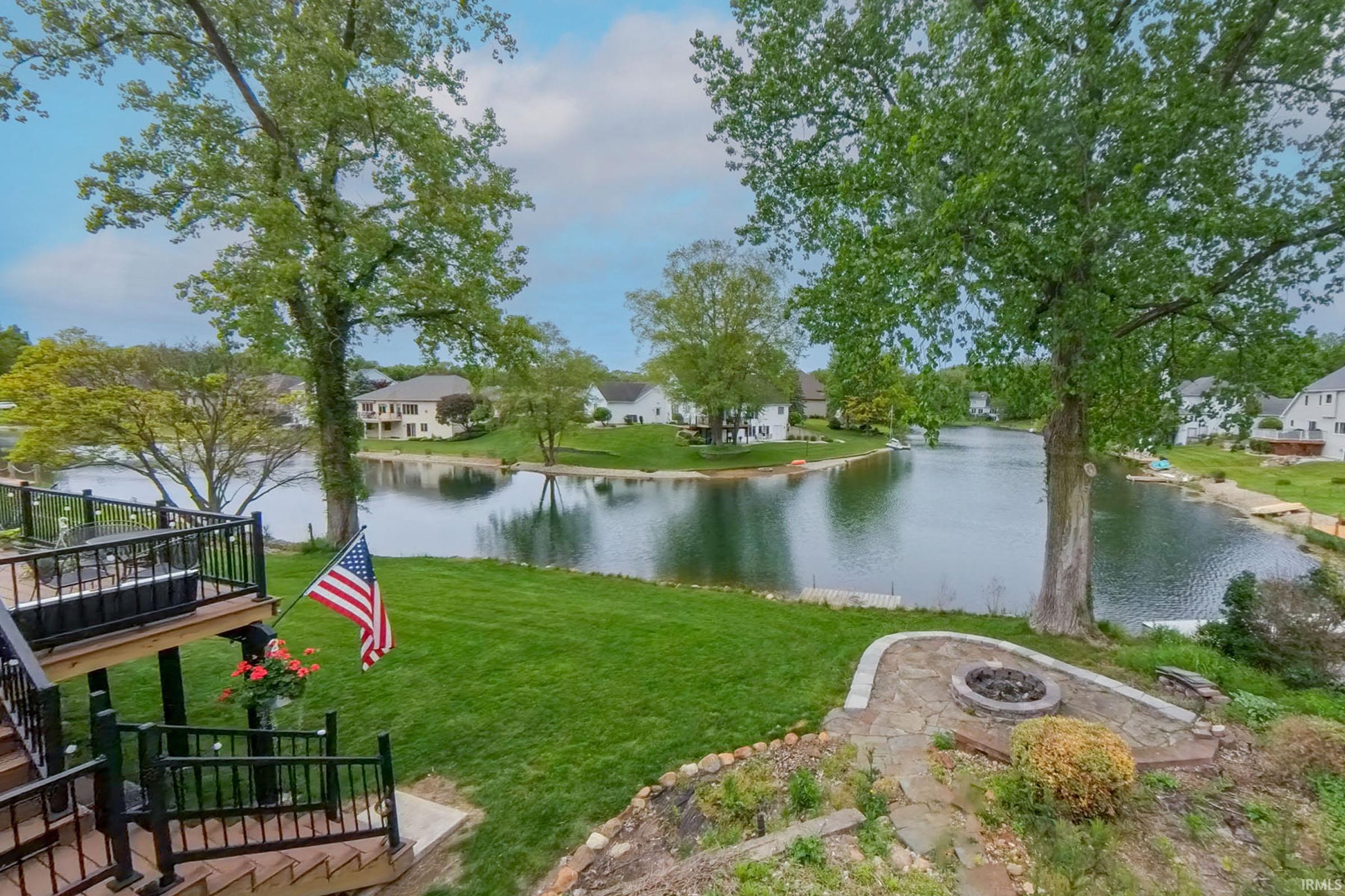 Expansive lakeside property featuring a multi-level deck with black railings, a stone patio with a built-in fire pit, mature trees, and a manicured lawn extending to the water's edge