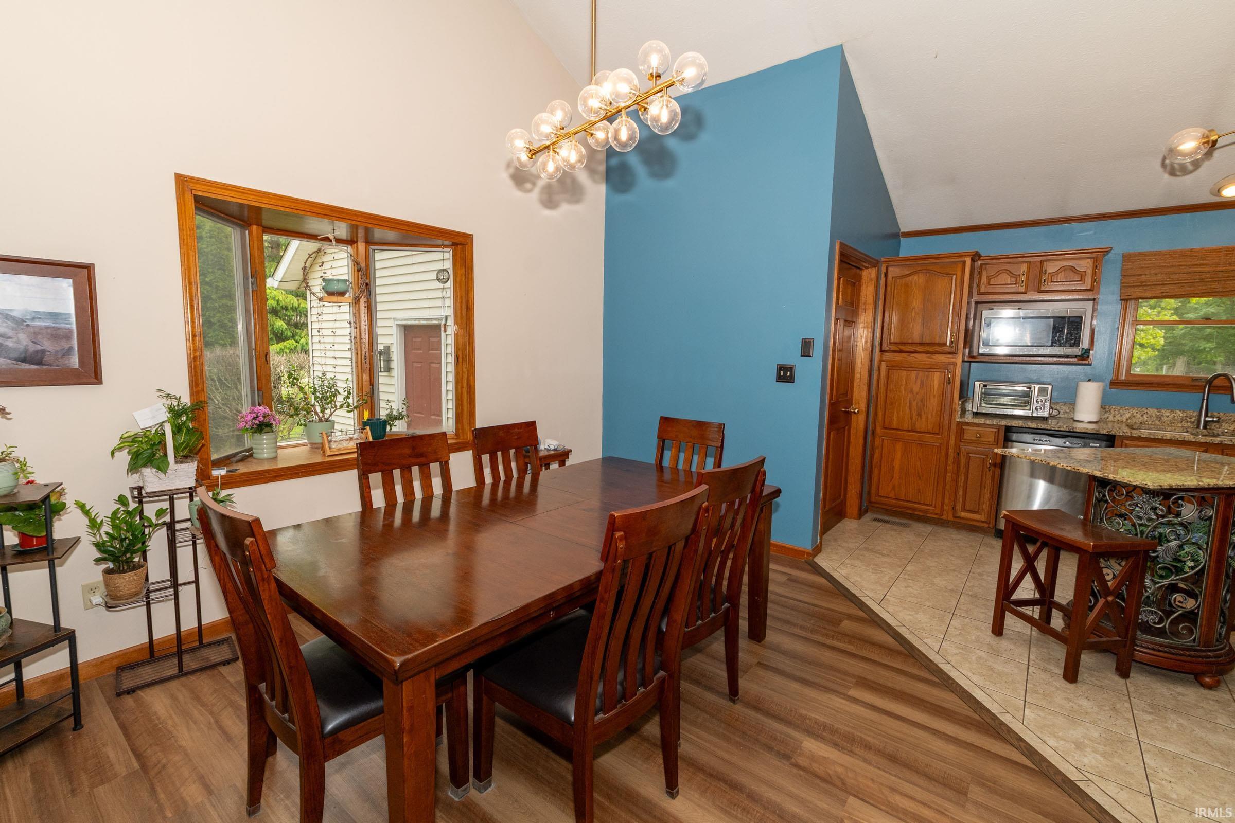 Open-concept dining area featuring vaulted ceilings, wood-finish flooring, a large bay window with wood trim, and a contemporary linear chandelier